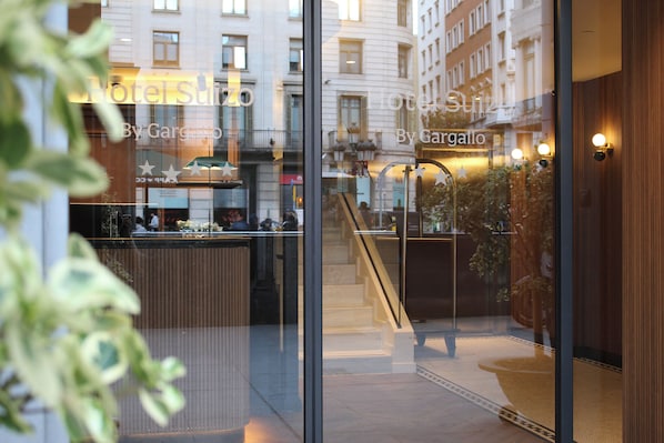 A hotel interior with a reception desk, a staircase, and a view of the street outside.