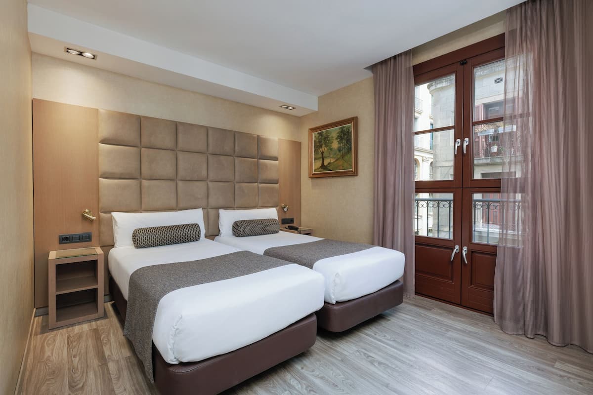 Ein Hotelzimmer mit zwei Betten, einem hölzernen Kopfteil, einem Nachttisch, einem Gemälde und einem großen Fenster mit Vorhängen.