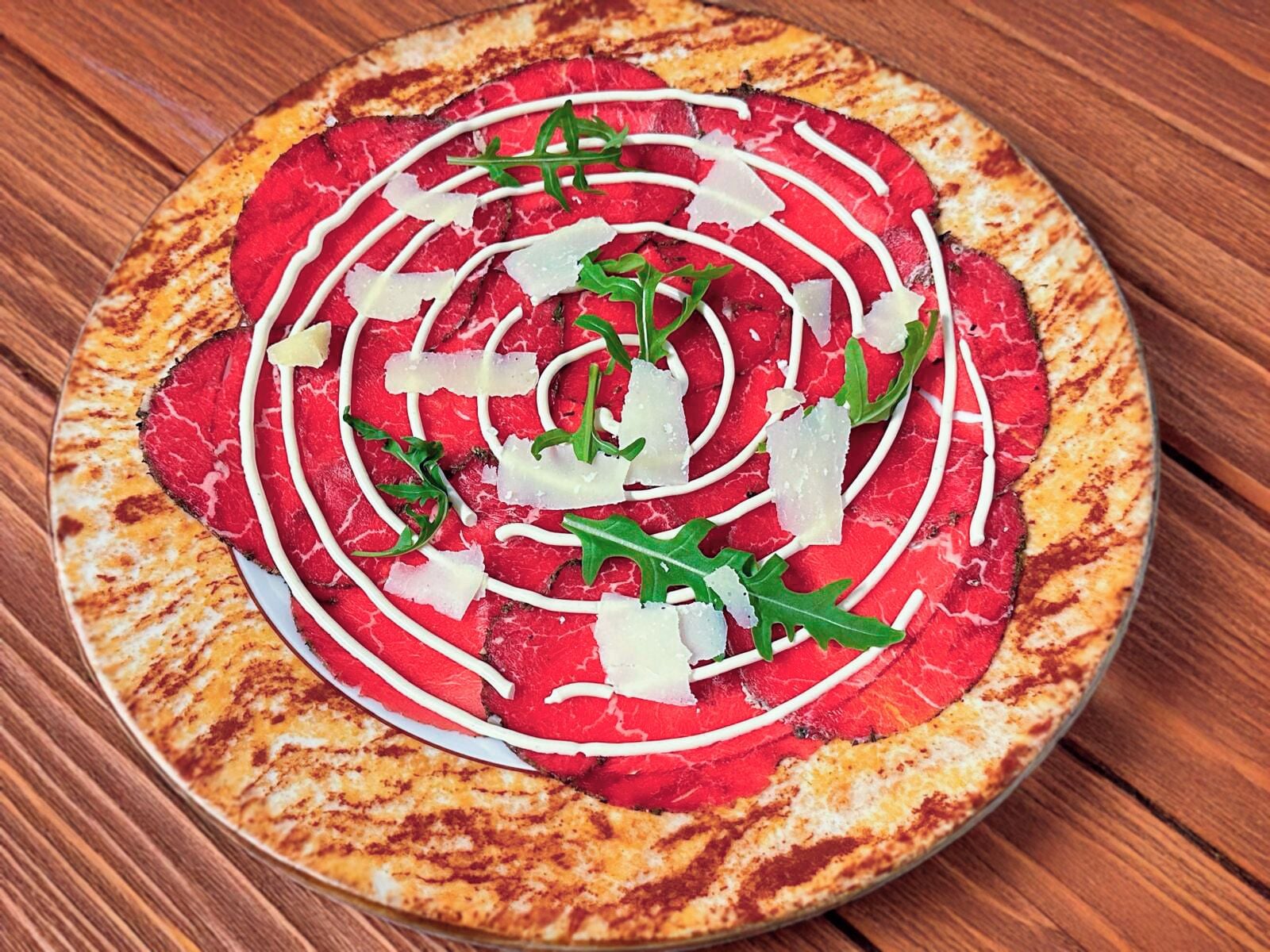 A pizza with a charred crust, topped with red meat, white sauce, cheese, and greens.