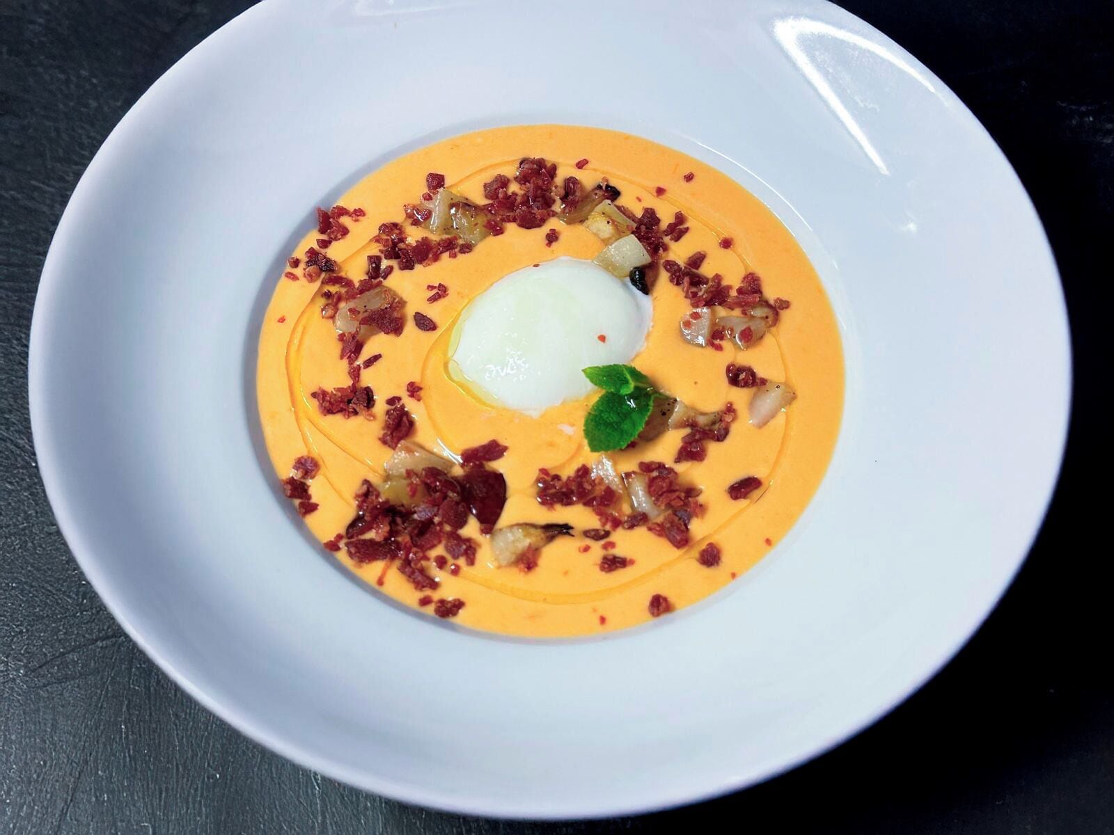 A white plate with a creamy orange soup, topped with a poached egg, crispy bacon bits, and a green leaf garnish.