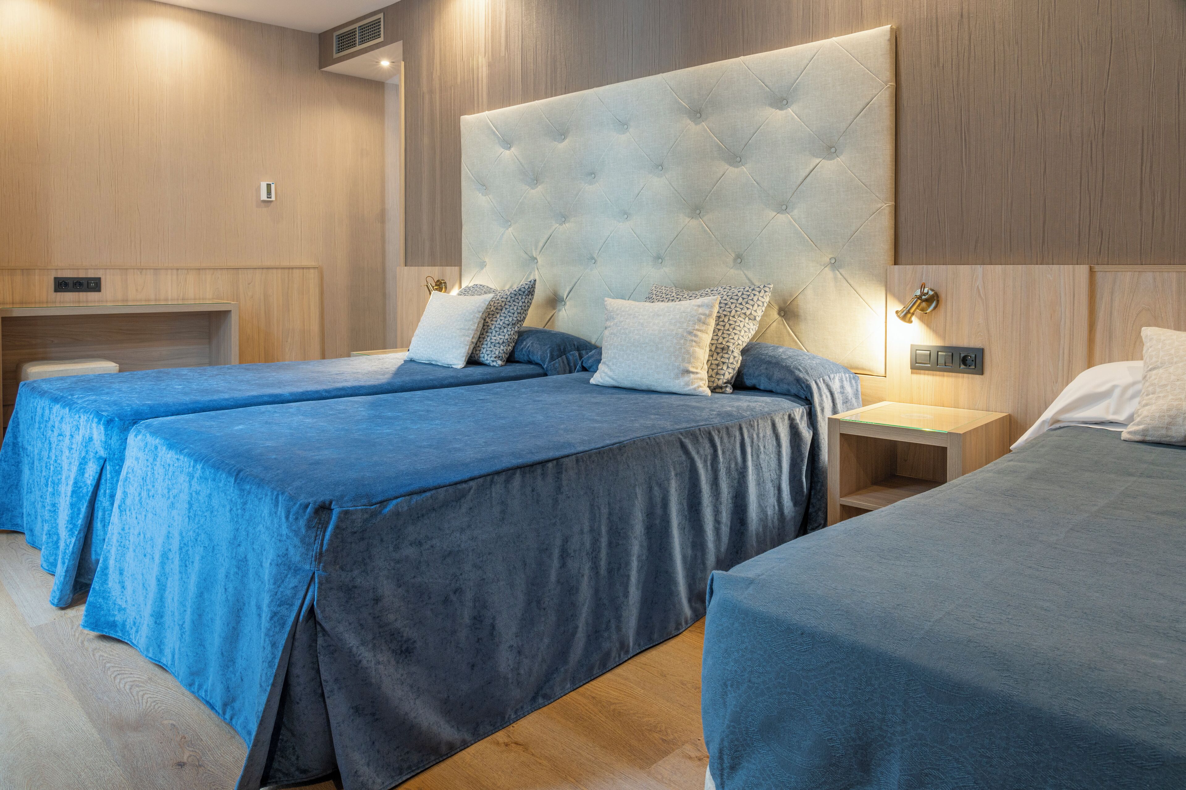 A hotel room with two beds, each with a blue bedspread and matching pillows. There is a wooden headboard with a tufted design, and a small wooden nightstand with a lamp.