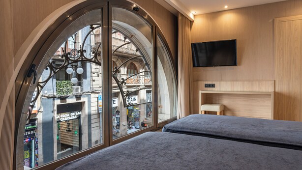 A hotel room with two beds, a large arched window, a mounted TV, and a small bedside table. - Rialto (Barcelona)