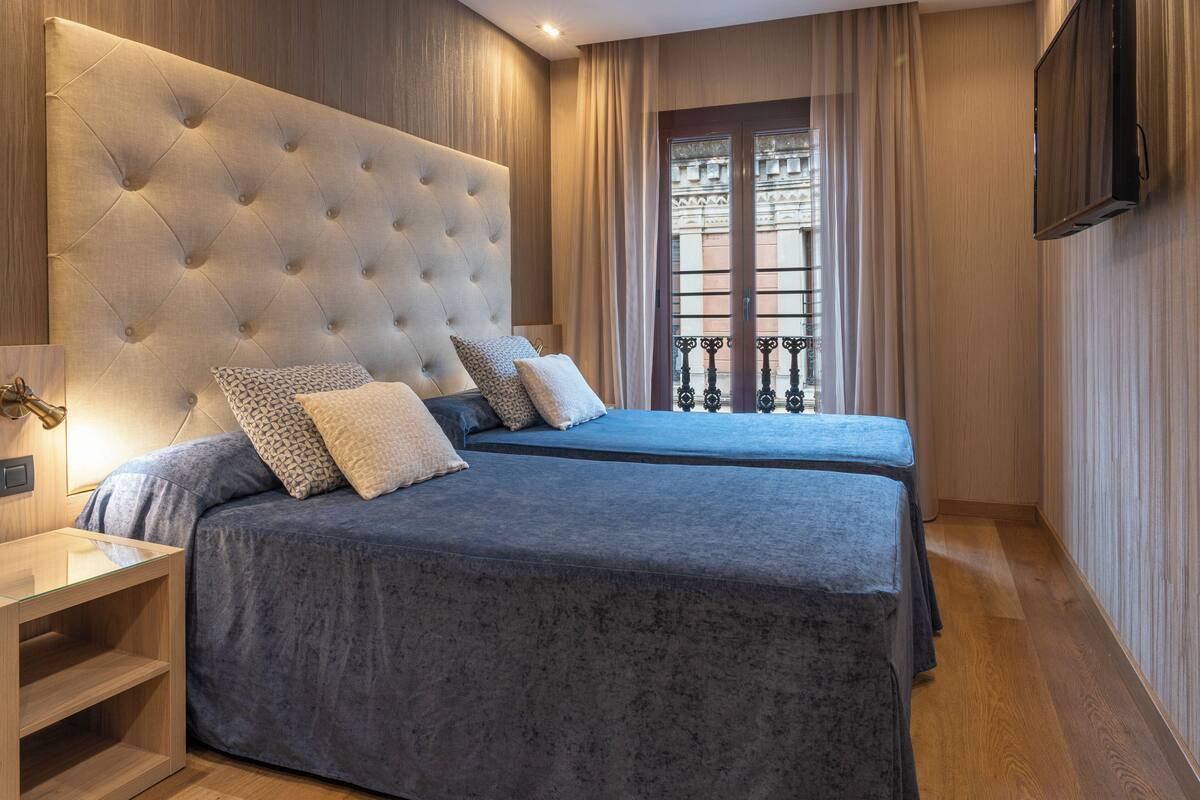 A bedroom with a tufted headboard, two beds with matching blue bedding, a bedside table, a TV mounted on the wall, and a window with curtains.