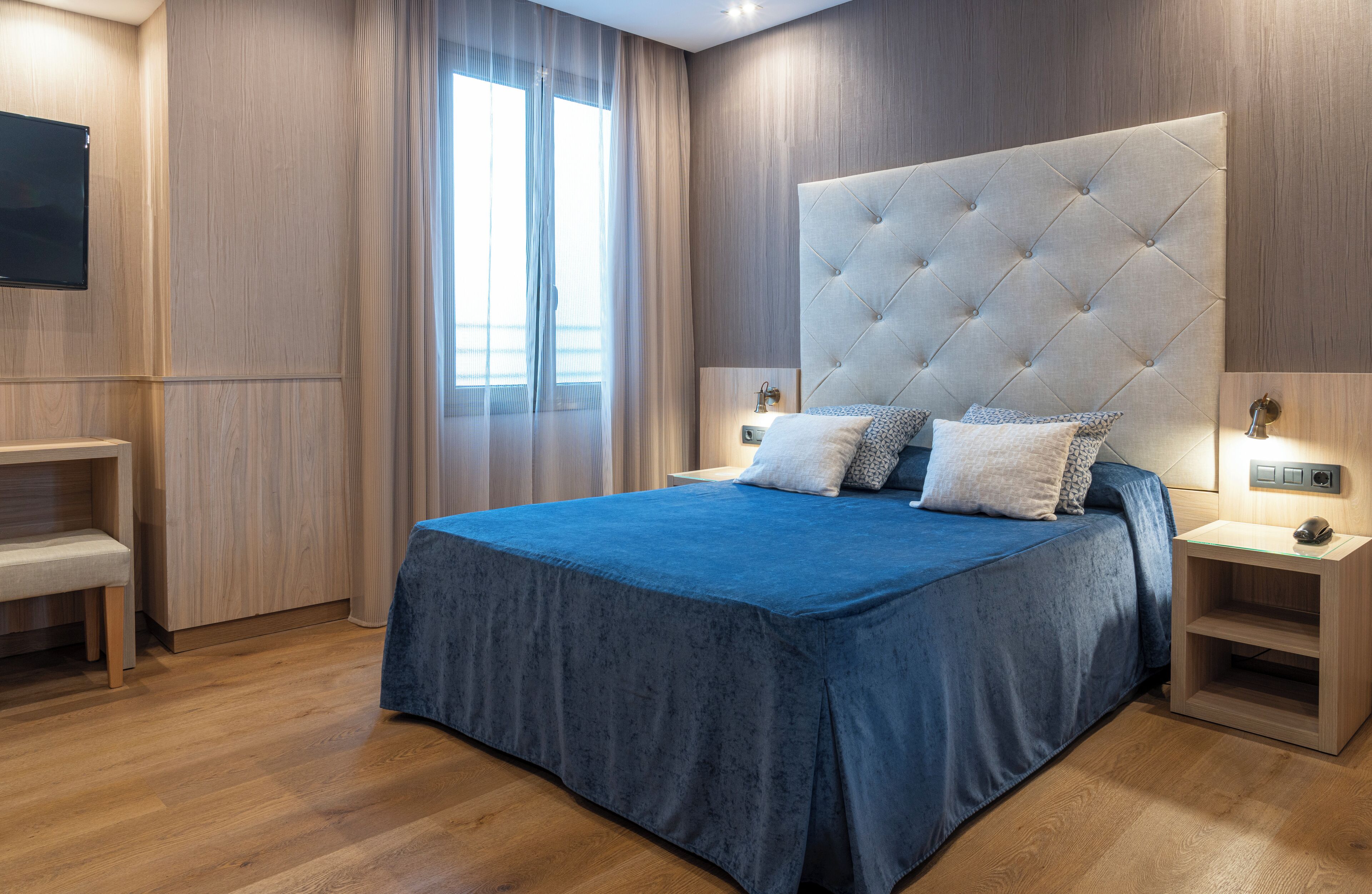 A modern bedroom with a large bed, wooden flooring, a TV mounted on the wall, and a bedside table with a lamp.