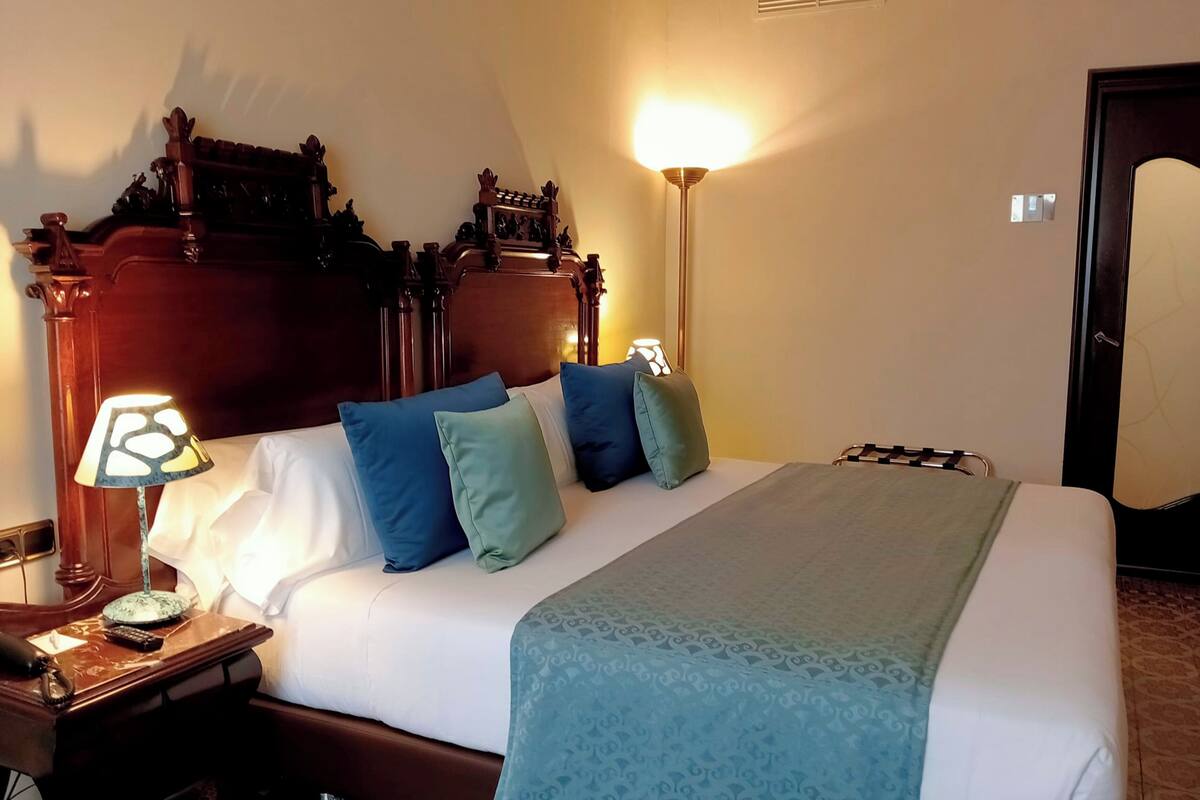 A hotel room with a large bed, a wooden headboard, a nightstand with a lamp, and a door.