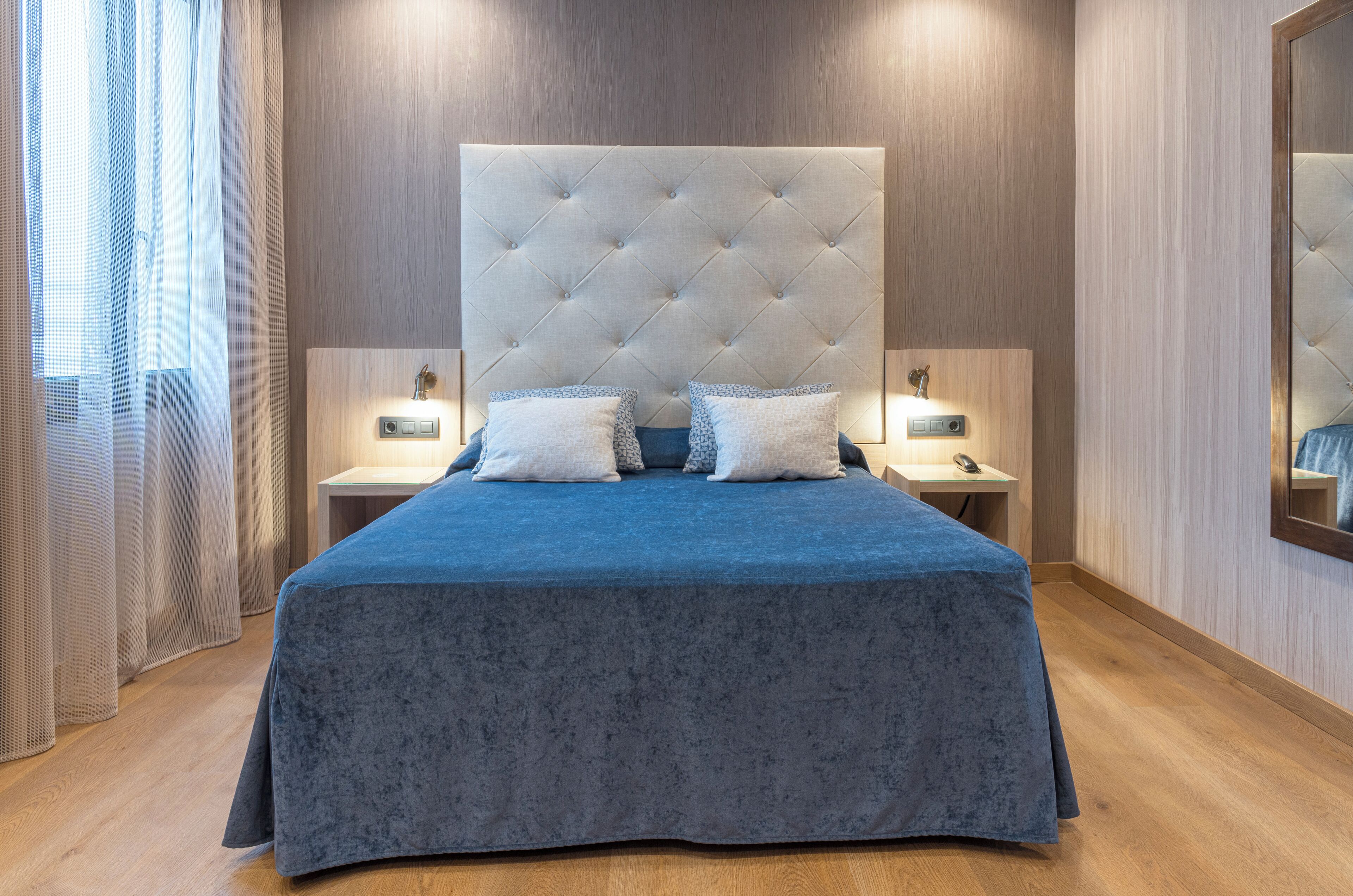 Una camera da letto moderna con una testiera trapuntata, una coperta blu, due comodini con lampade e un grande specchio a parete.