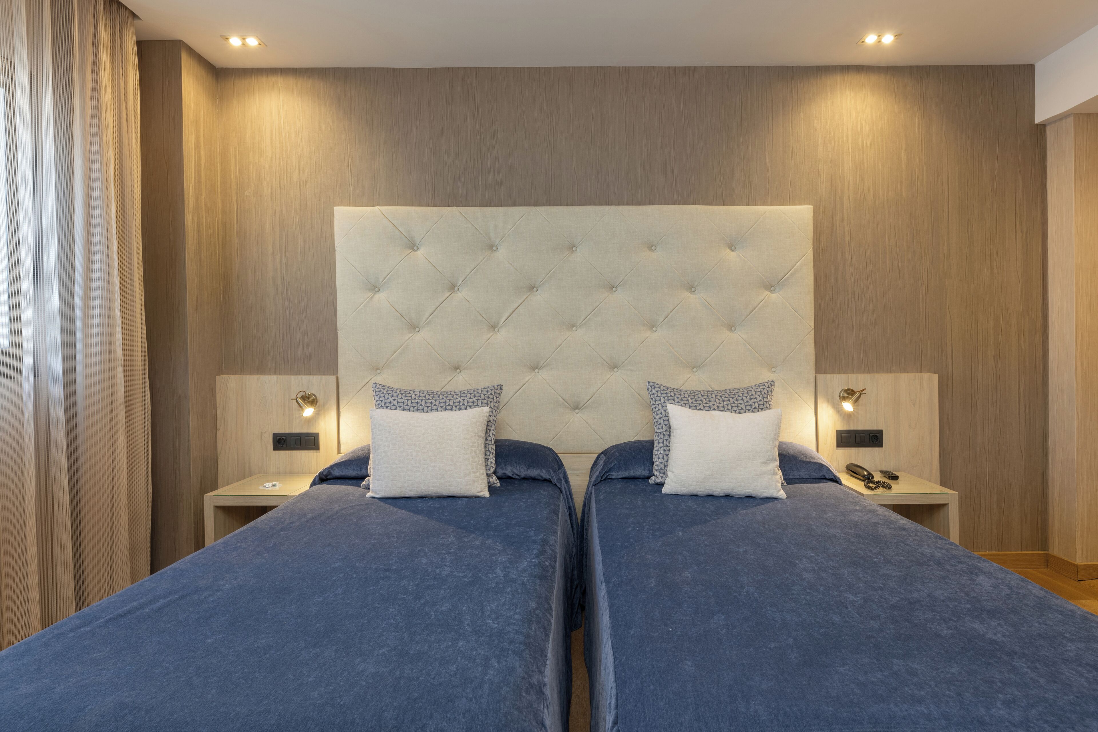 Une chambre moderne avec deux lits, une grande tête de lit capitonnée, des lampes de chevet et une table de chevet avec un téléphone.