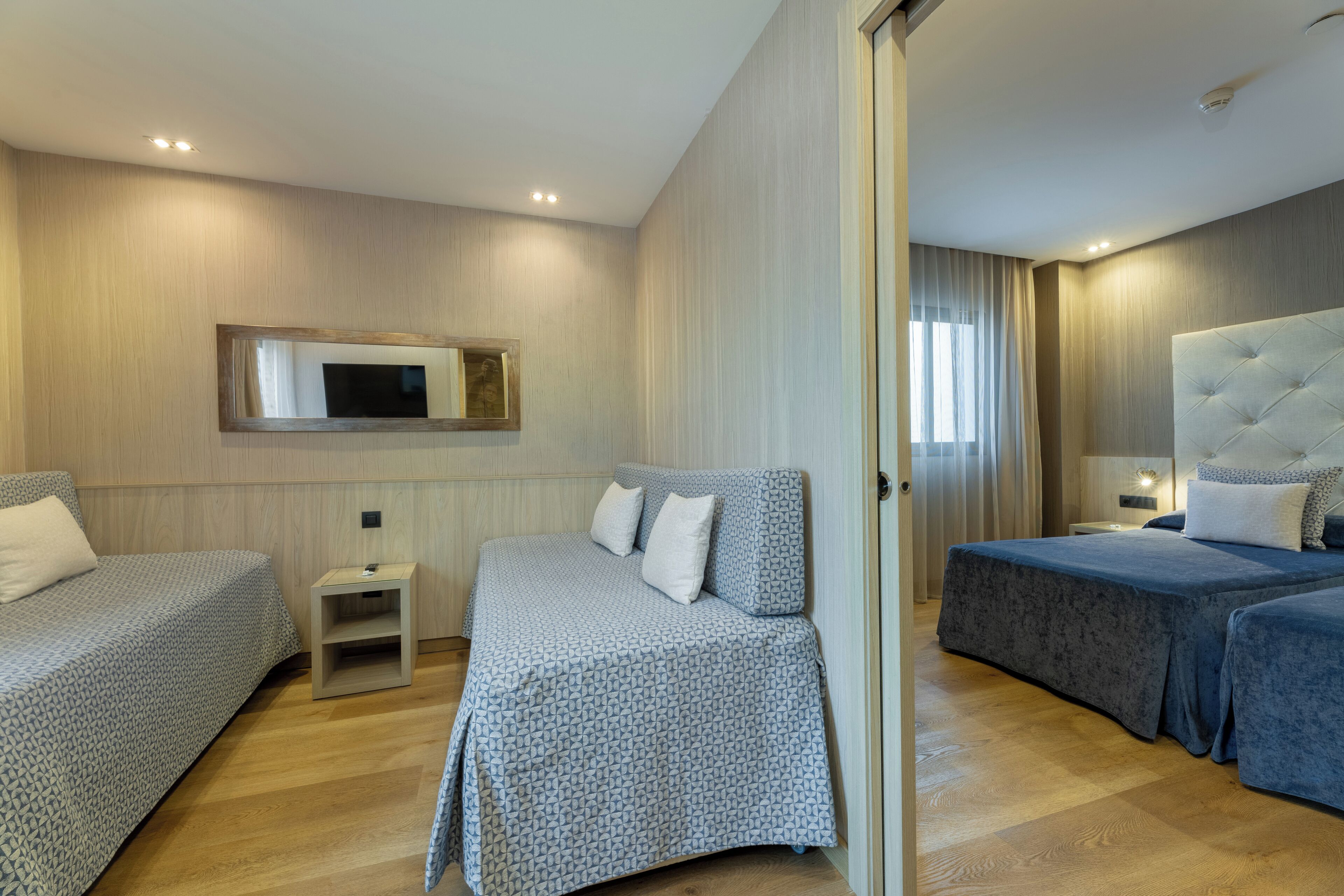 A hotel room with two single beds, a small bedside table, a large mirror, and a television on the wall.
