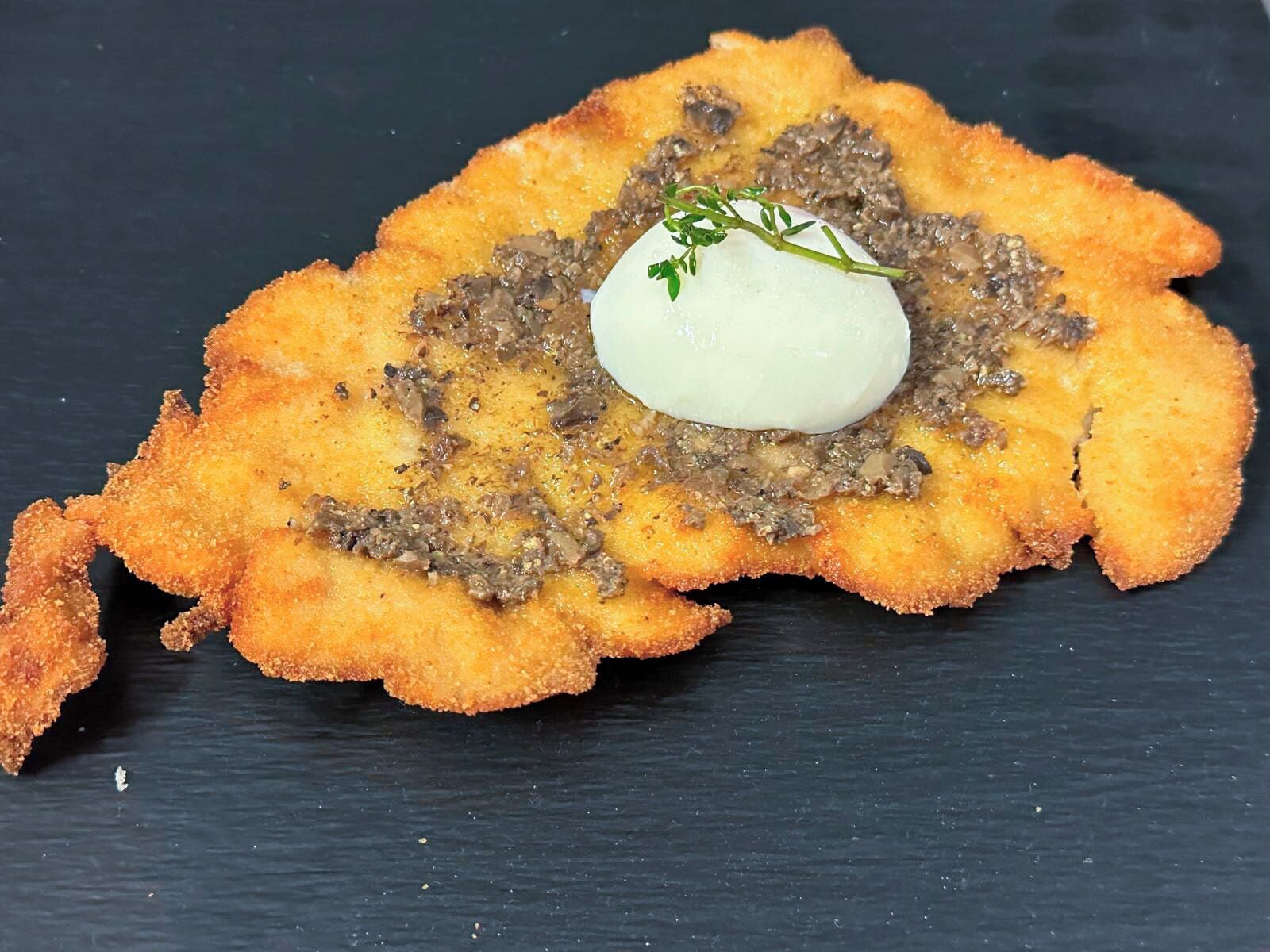 A dish with a crispy, golden-brown fried item, a dollop of white sauce, and a garnish of green herbs on a dark slate plate.