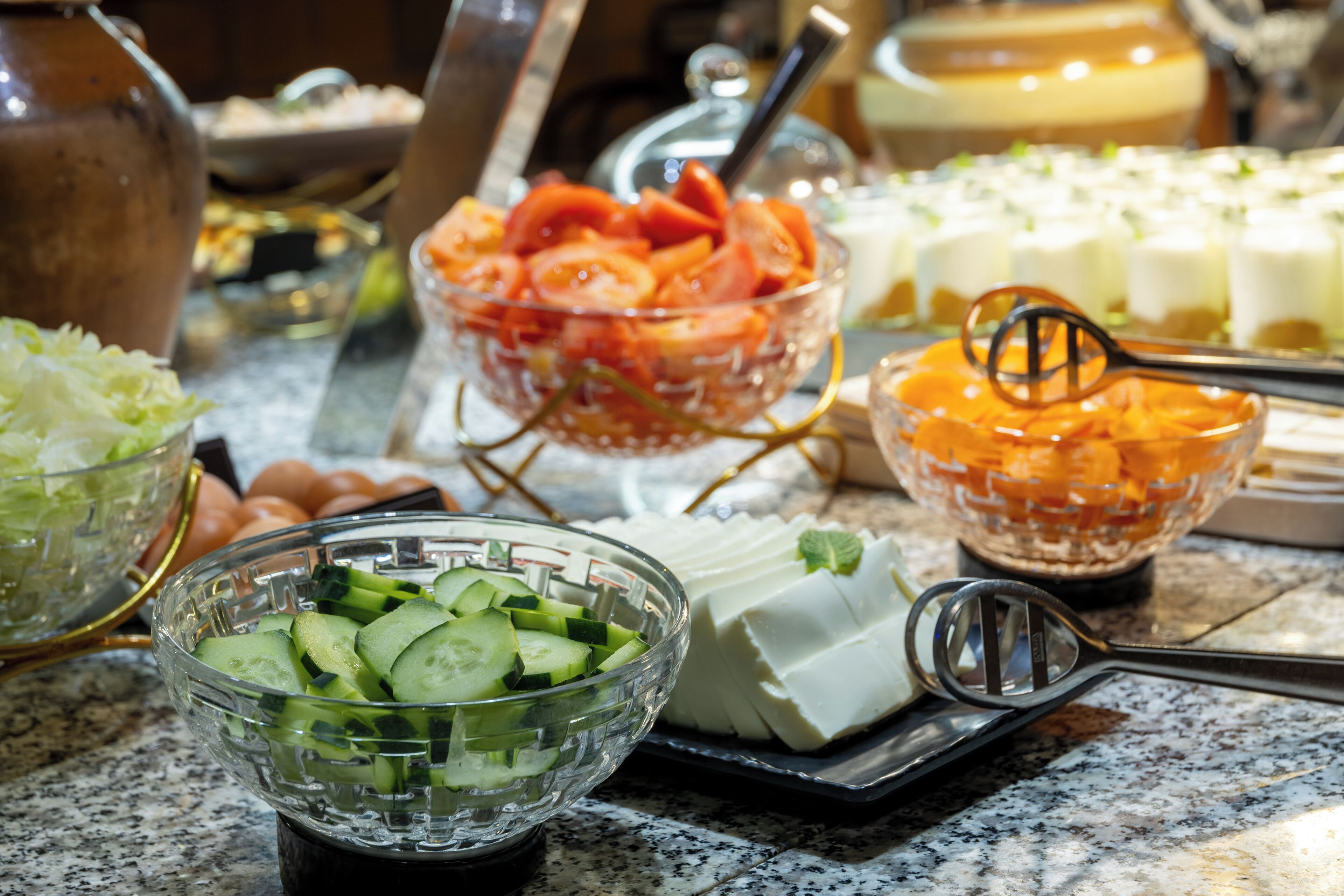 A kitchen counter with various food items including a bowl of sliced cucumbers, a bowl of sliced tomatoes, a bowl of sliced carrots, a plate of white cheese, and a bowl of dark sauce.