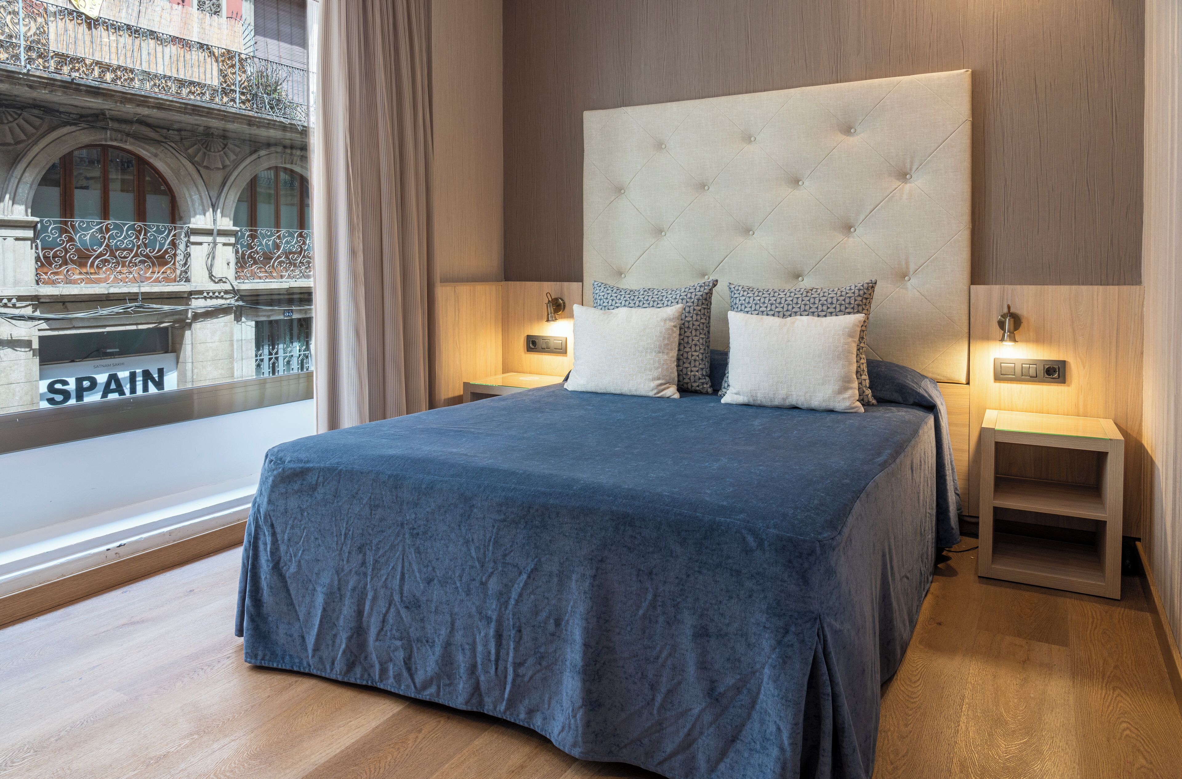 Una habitación de hotel moderna con una cama grande, dos mesitas de noche y vistas a un edificio con la palabra 'SPAIN' en la ventana.