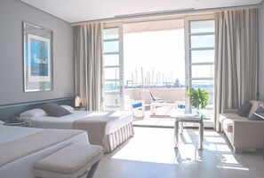 Una habitación de hotel con dos camas, un balcón con vistas al puerto deportivo y una zona de estar con mesa y sillas.