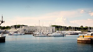 Un port de plaisance où sont amarrés de nombreux bateaux, un bâtiment moderne en arrière-plan et un ciel dégagé.