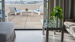 Un salon moderne avec une table en verre, deux fauteuils blancs et une vue sur une plage où se trouvent des bateaux.