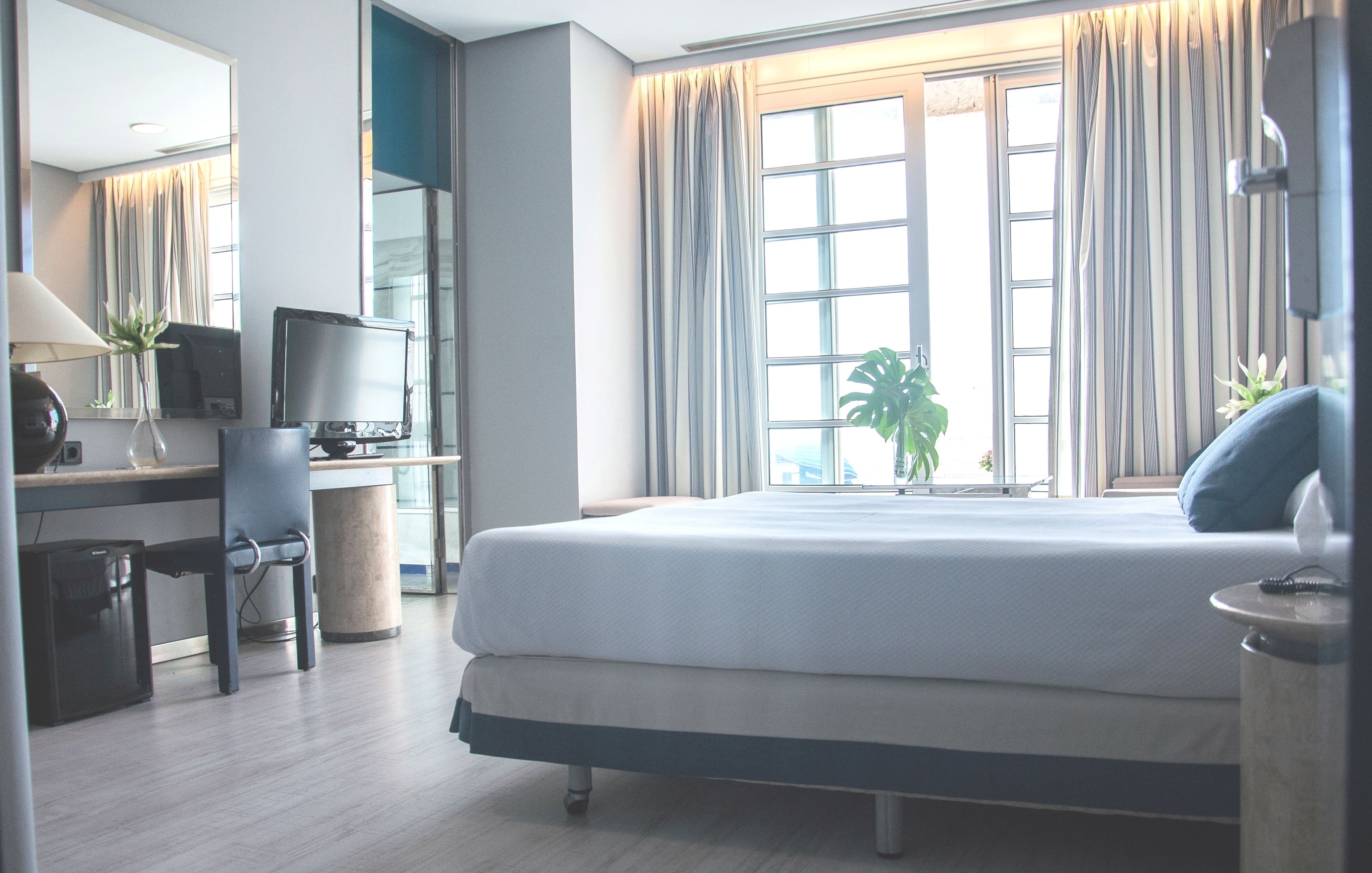 A hotel room with a bed, a desk with a computer, a chair, and a window with curtains.