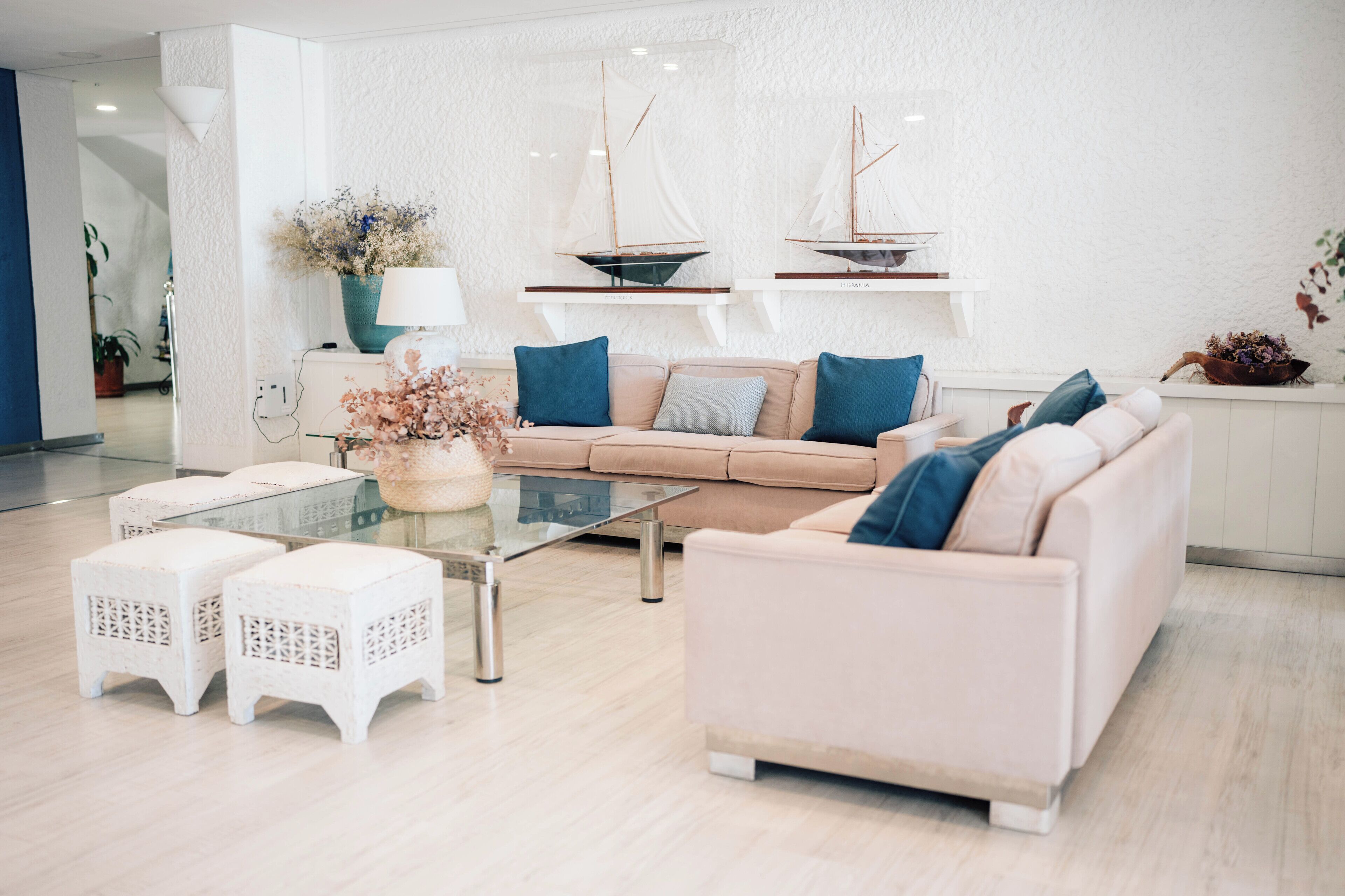 Een moderne woonkamer met een beige bank, twee witte zitkussens, een glazen salontafel en een witte muur met twee decoraties van zeilboten.