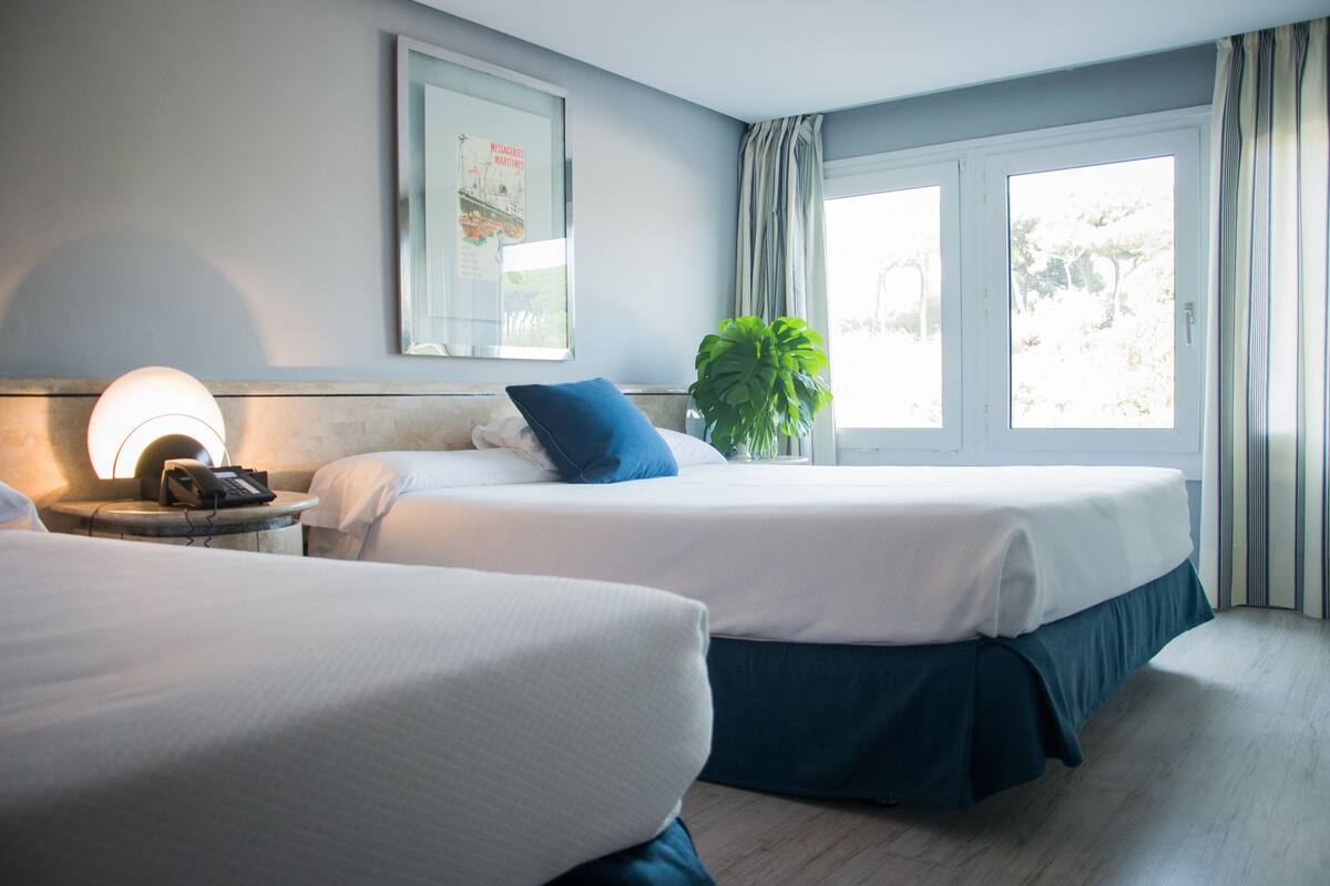 Habitación de hotel con dos camas, una mesita de noche con lámpara, un cuadro enmarcado en la pared y una ventana con cortinas.