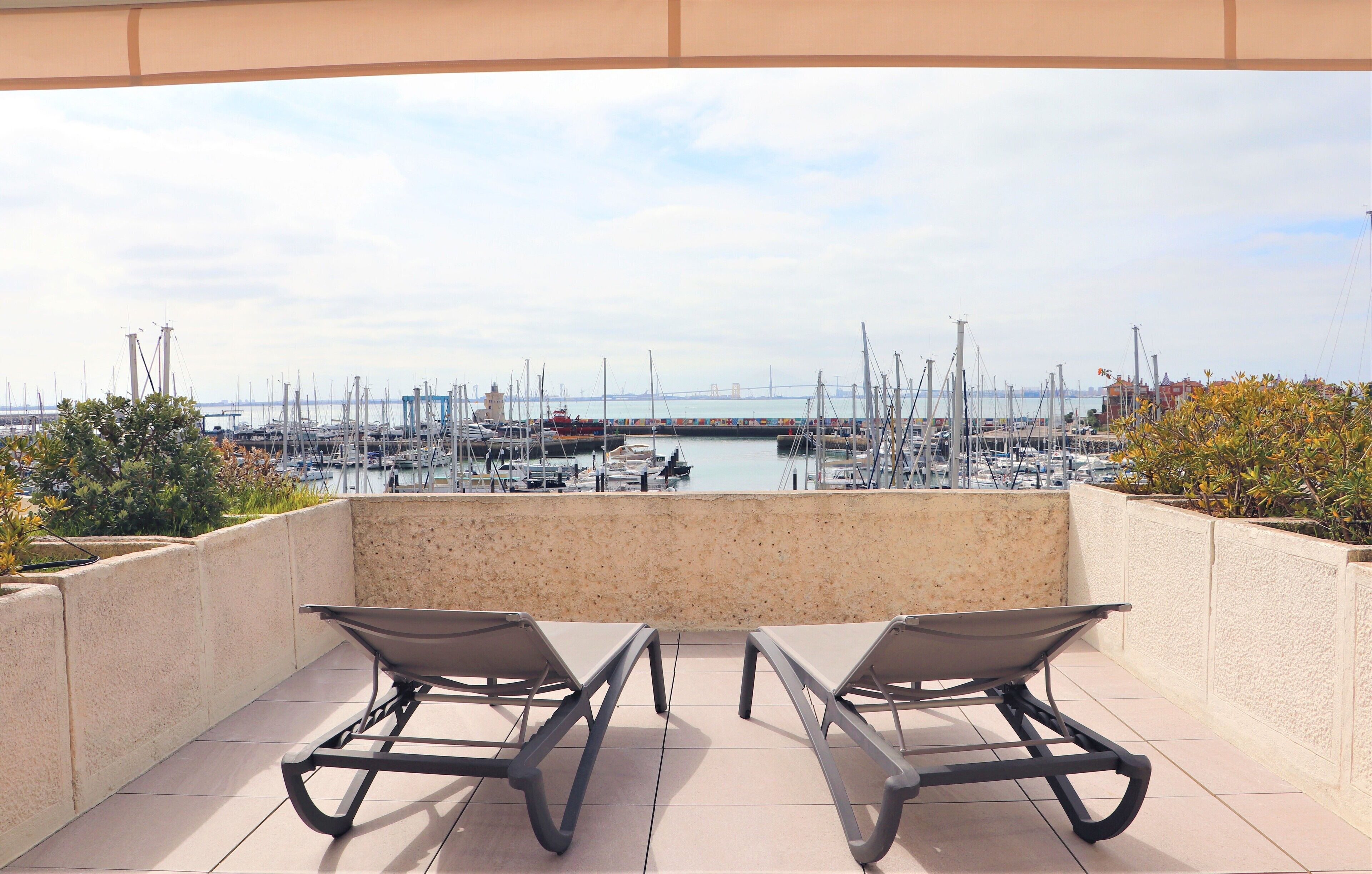Un balcon avec deux chaises longues donnant sur une marina où sont amarrés des bateaux, et où l’on aperçoit un pont au loin.