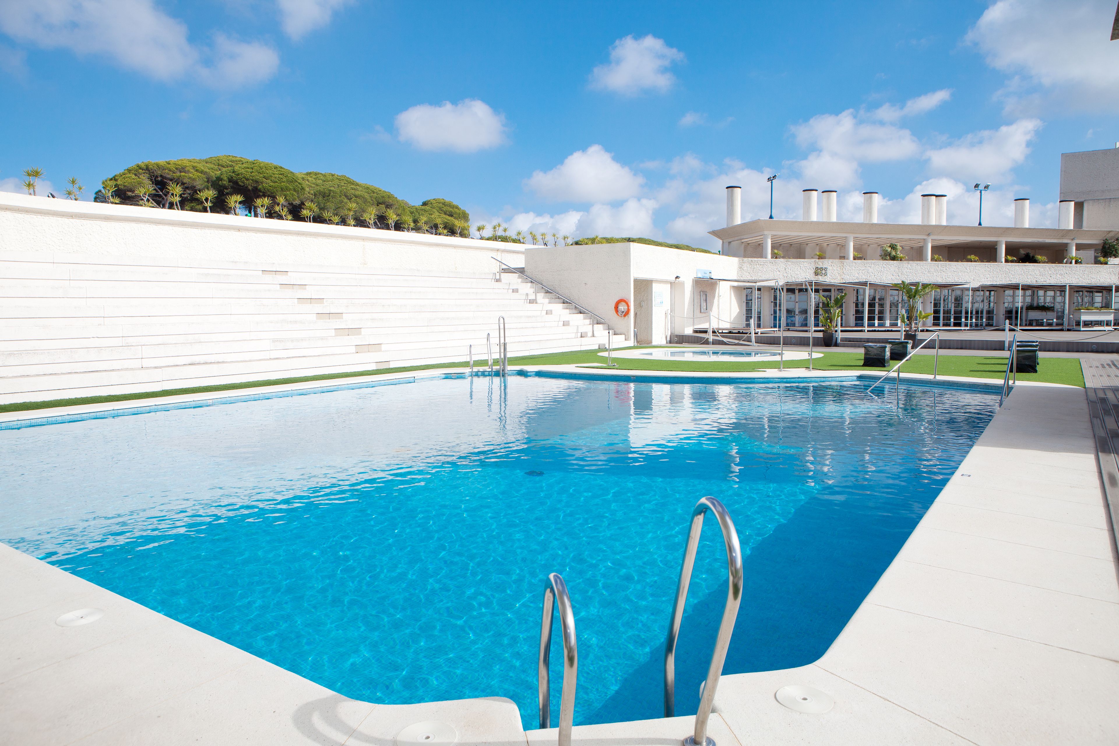 A swimming pool with clear blue water, a white poolside area, and a staircase leading to a building with a white facade.