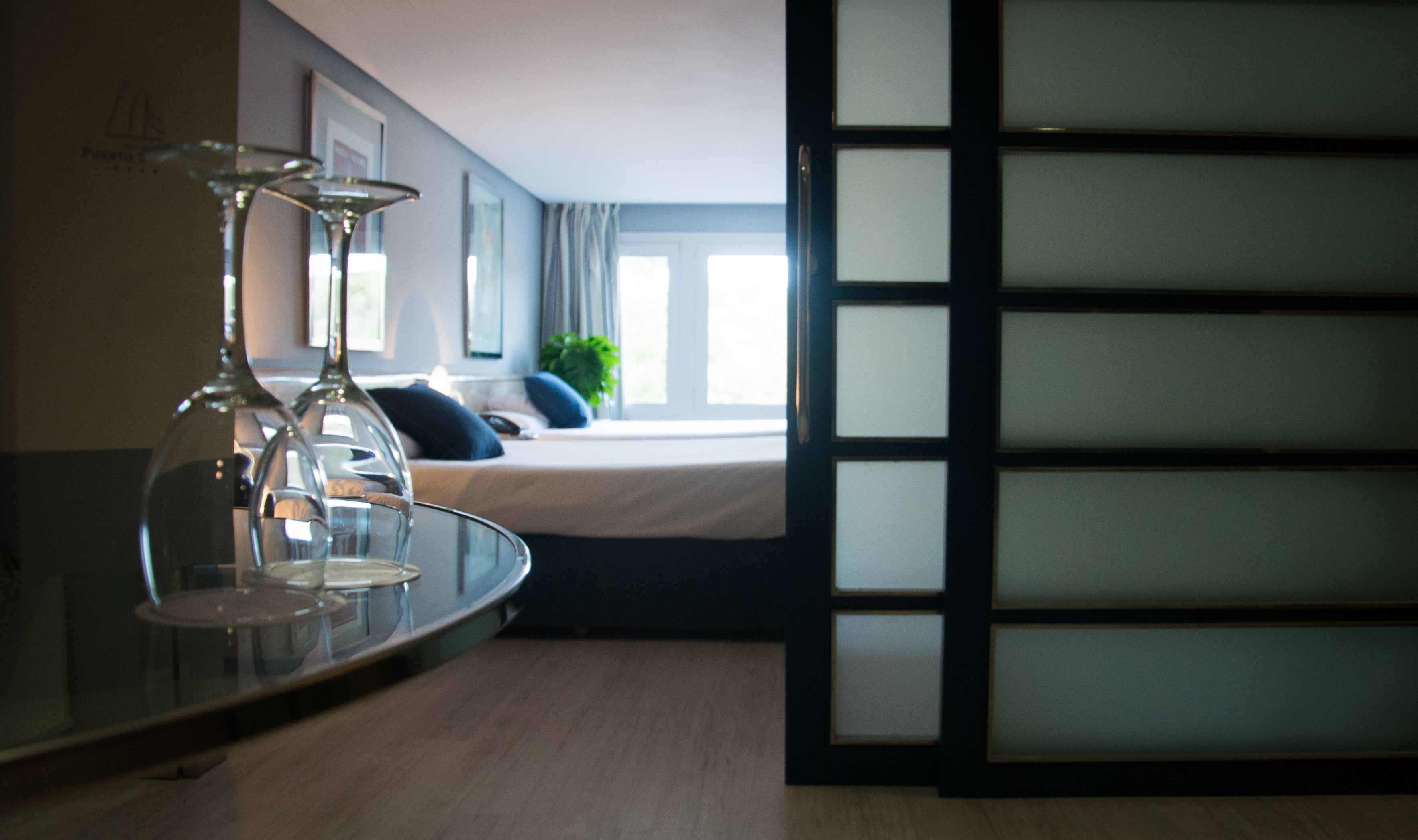 A hotel room with a bed, a glass table with a wine glass, and a view of the outdoors through a window.