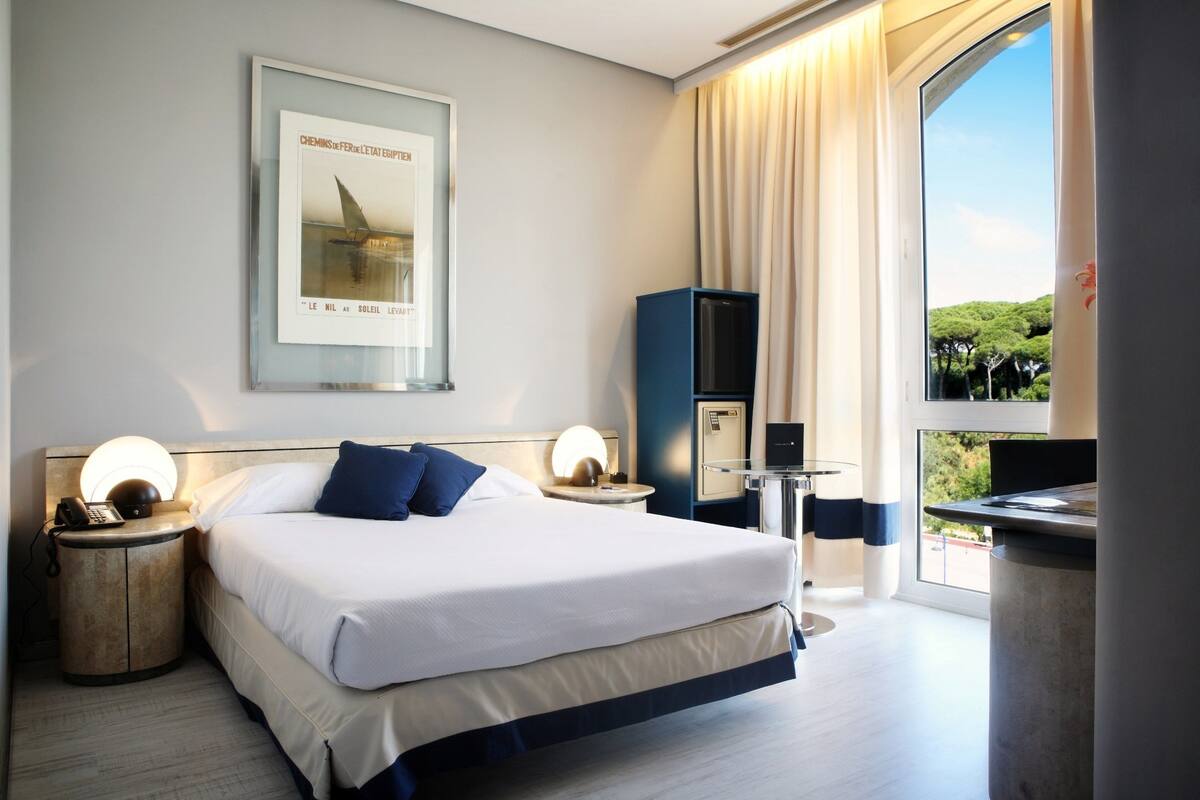 Habitación de hotel moderna con una cama grande, lámparas de noche, un escritorio con una computadora y un ventanal con vistas a zonas verdes.