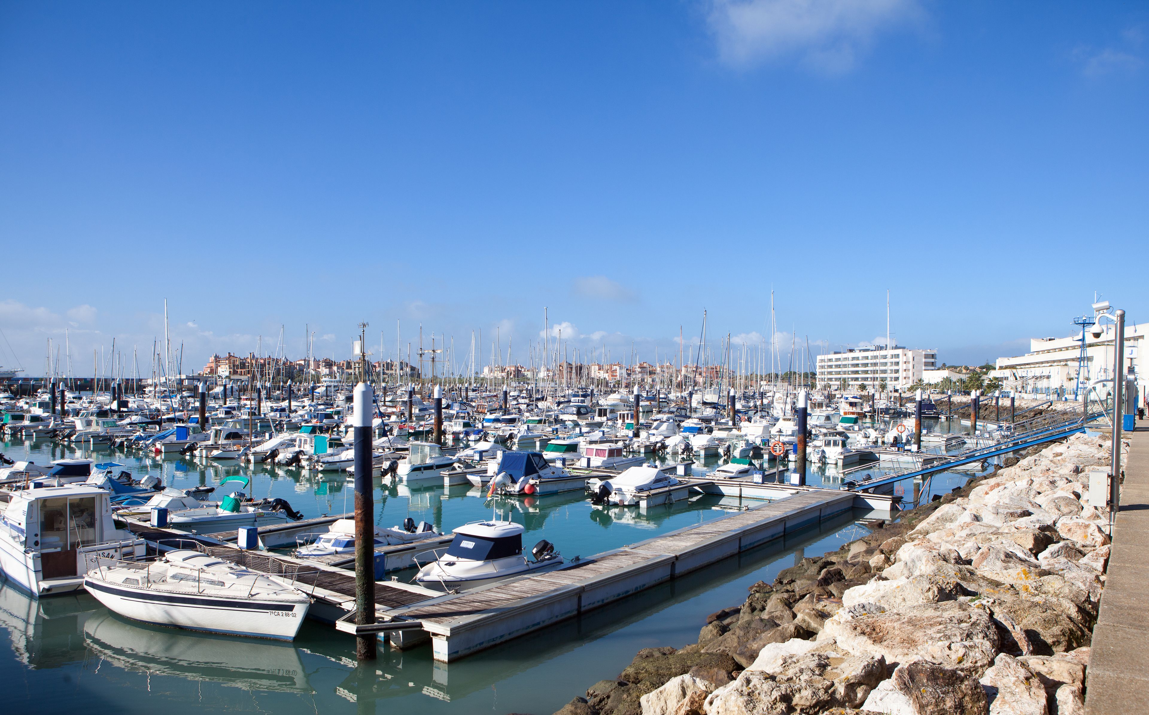 A marina with numerous boats docked at wooden piers, a rocky shoreline, and a clear blue sky.