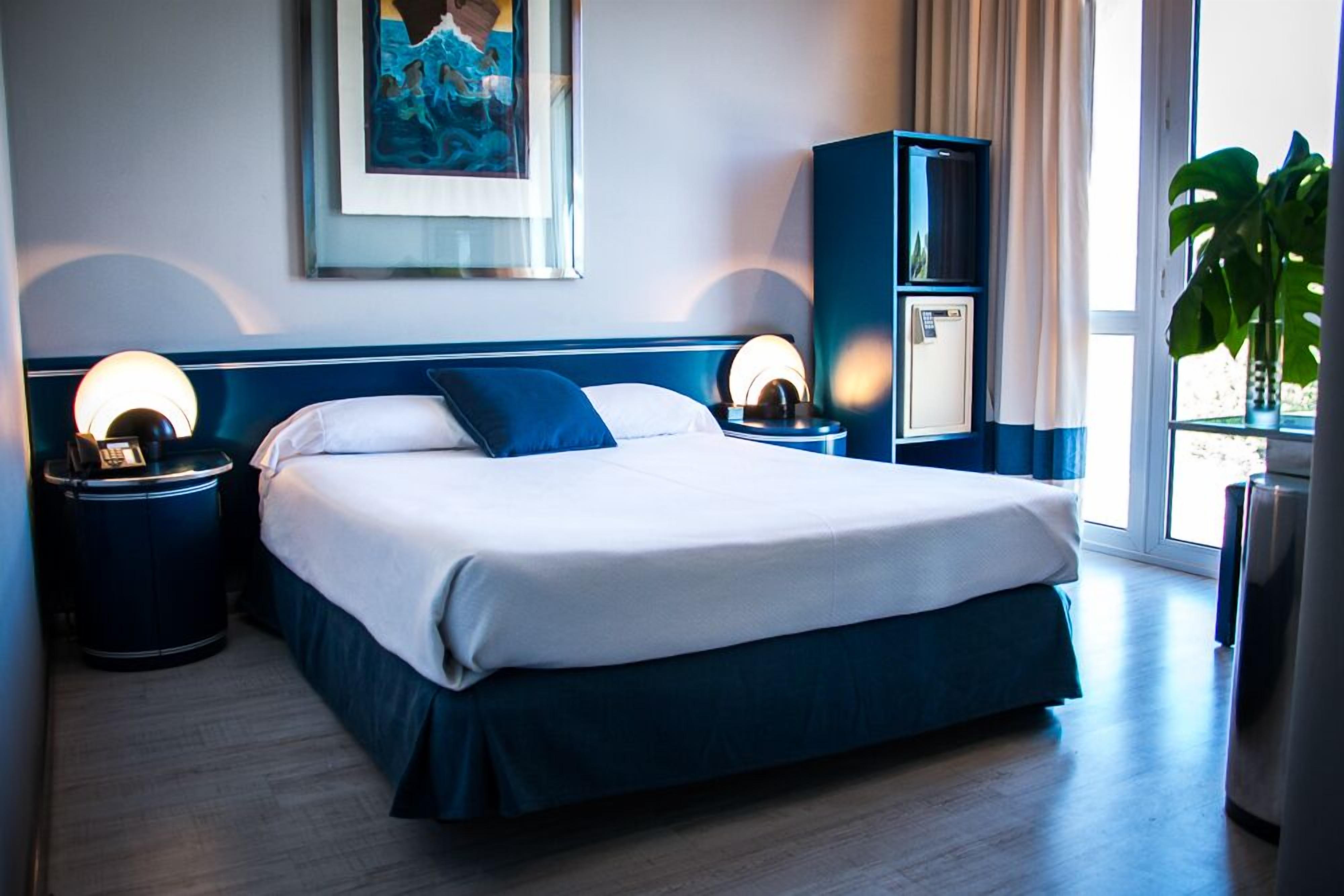 Une chambre d’hôtel avec un grand lit, deux tables de chevet avec des lampes, un minibar et une grande fenêtre avec des rideaux.