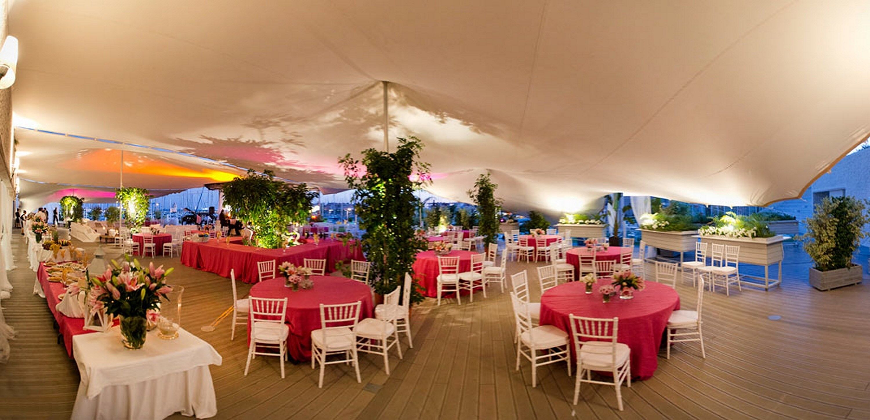 Un espacio para eventos al aire libre con mesas redondas cubiertas con manteles rojos y sillas blancas, bajo un gran toldo blanco.
