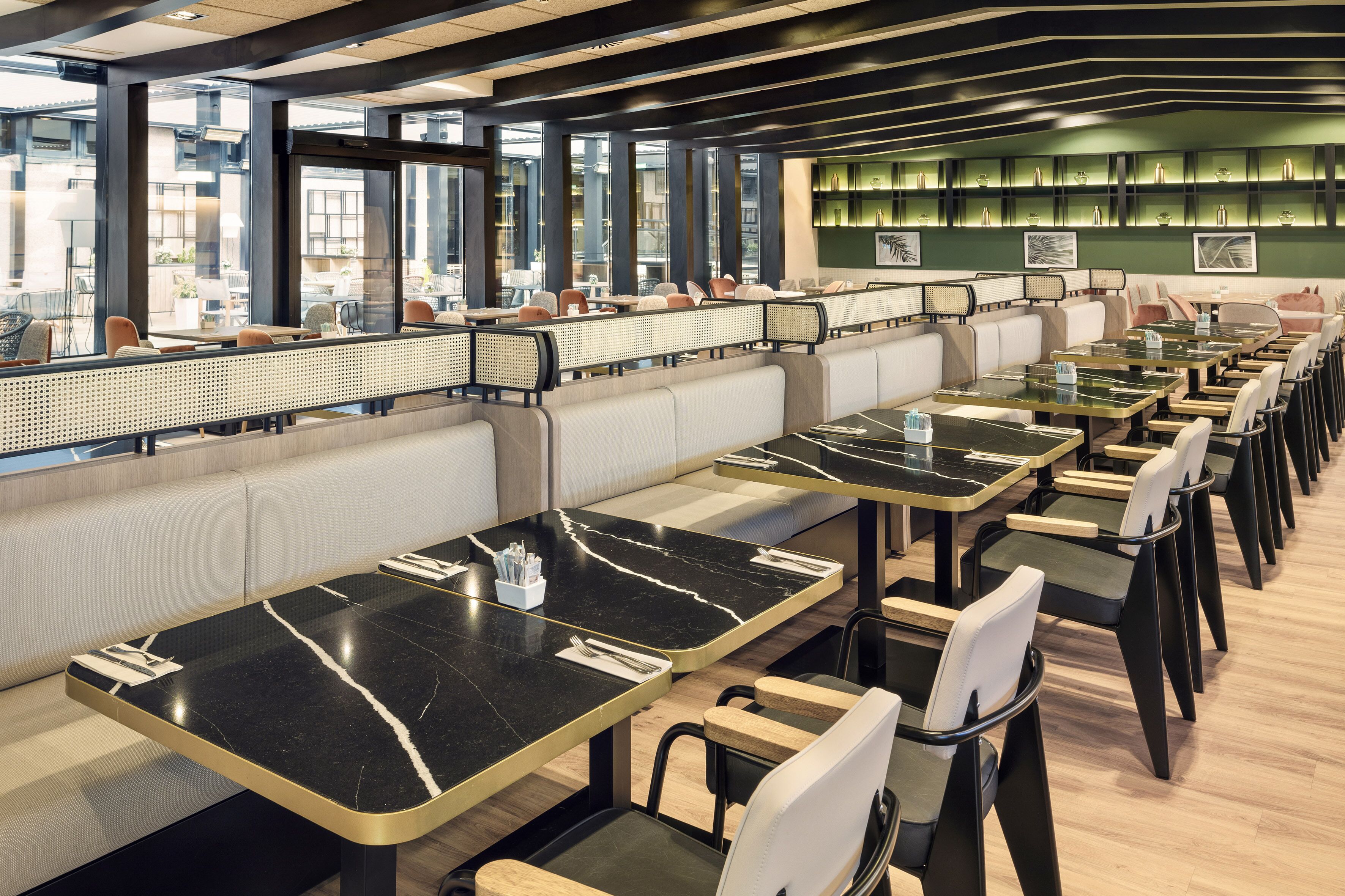 Un restaurante moderno con mesas comunitarias largas y taburetes altos, ventanas amplias y una pared verde decorativa con estanterías.