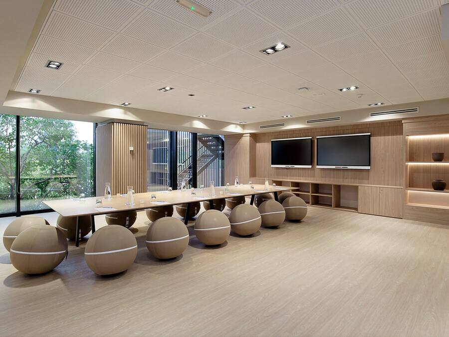 A modern conference room with a long table, round seating, a large TV, and a view of greenery outside.