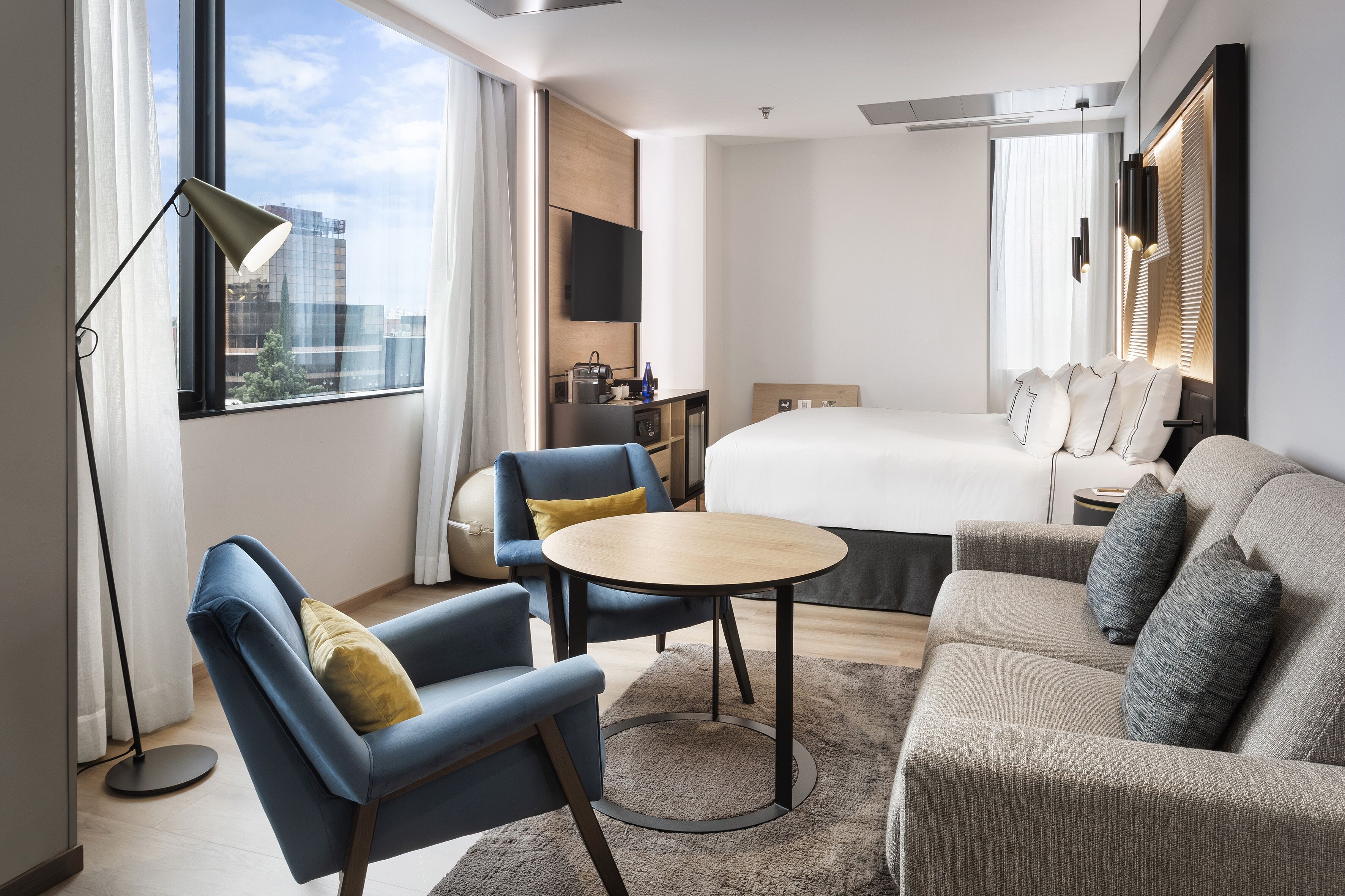 Une chambre d’hôtel moderne équipée d’un lit, d’un canapé, d’un fauteuil, d’une petite table, d’une télévision et d’un bureau avec une lampe.