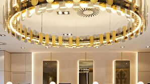 A modern reception area with a circular chandelier, wooden reception desks, and a ceiling with a geometric pattern. - Melia Avenida América (Madrid)