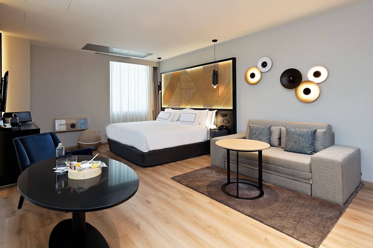 A modern hotel room with a large bed, a sofa, a round table, and a desk with a chair.