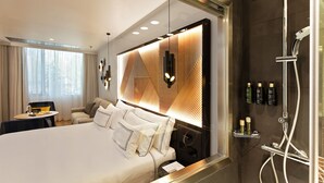 A modern hotel room with a large bed, a bedside table, a seating area with a chair and table, and a shower area with a handheld showerhead. - Melia Avenida América (Madrid)
