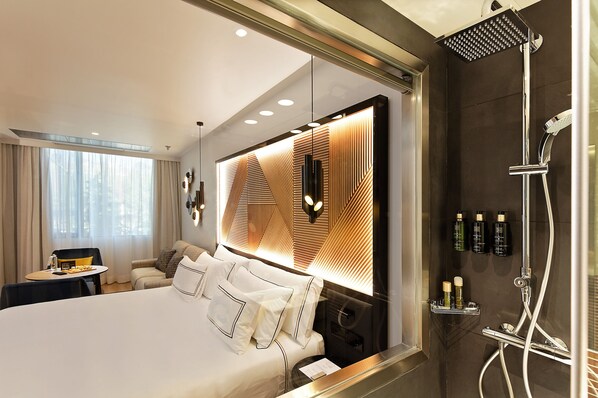 A modern hotel room with a large bed, a bedside table, a seating area with a chair and table, and a shower area with a handheld showerhead. - Melia Avenida América (Madrid)