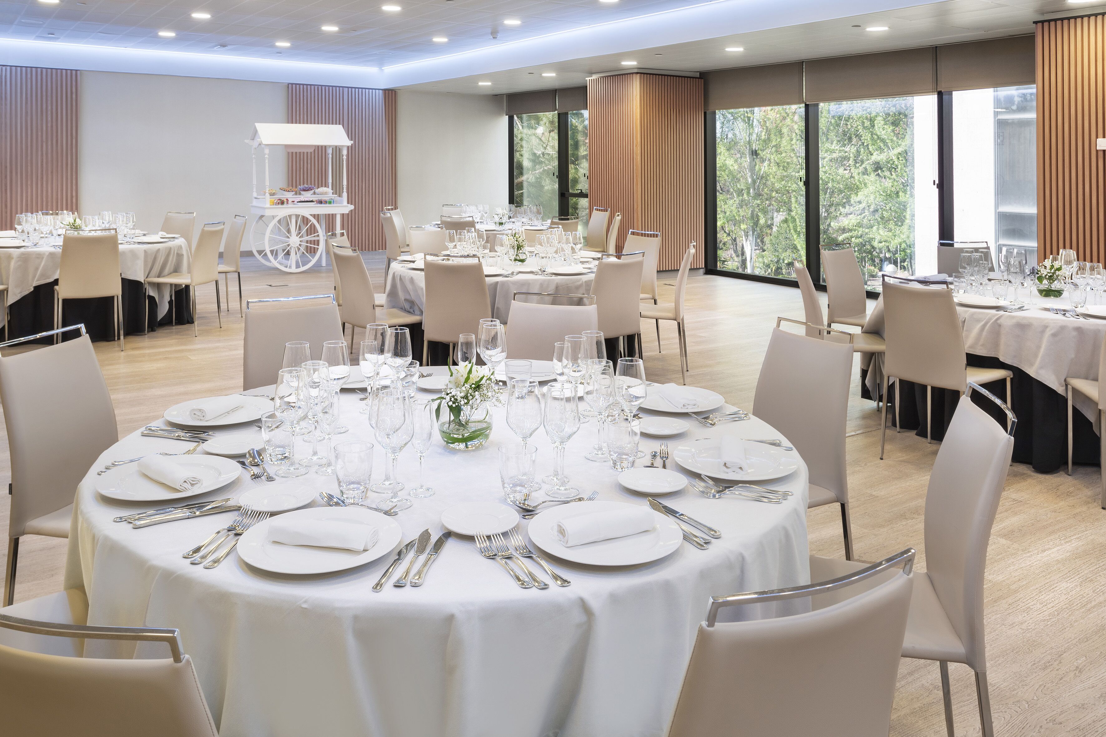 Uma sala de banquetes com mesas redondas preparadas para refeições, grandes janelas e um carrinho branco ao fundo.