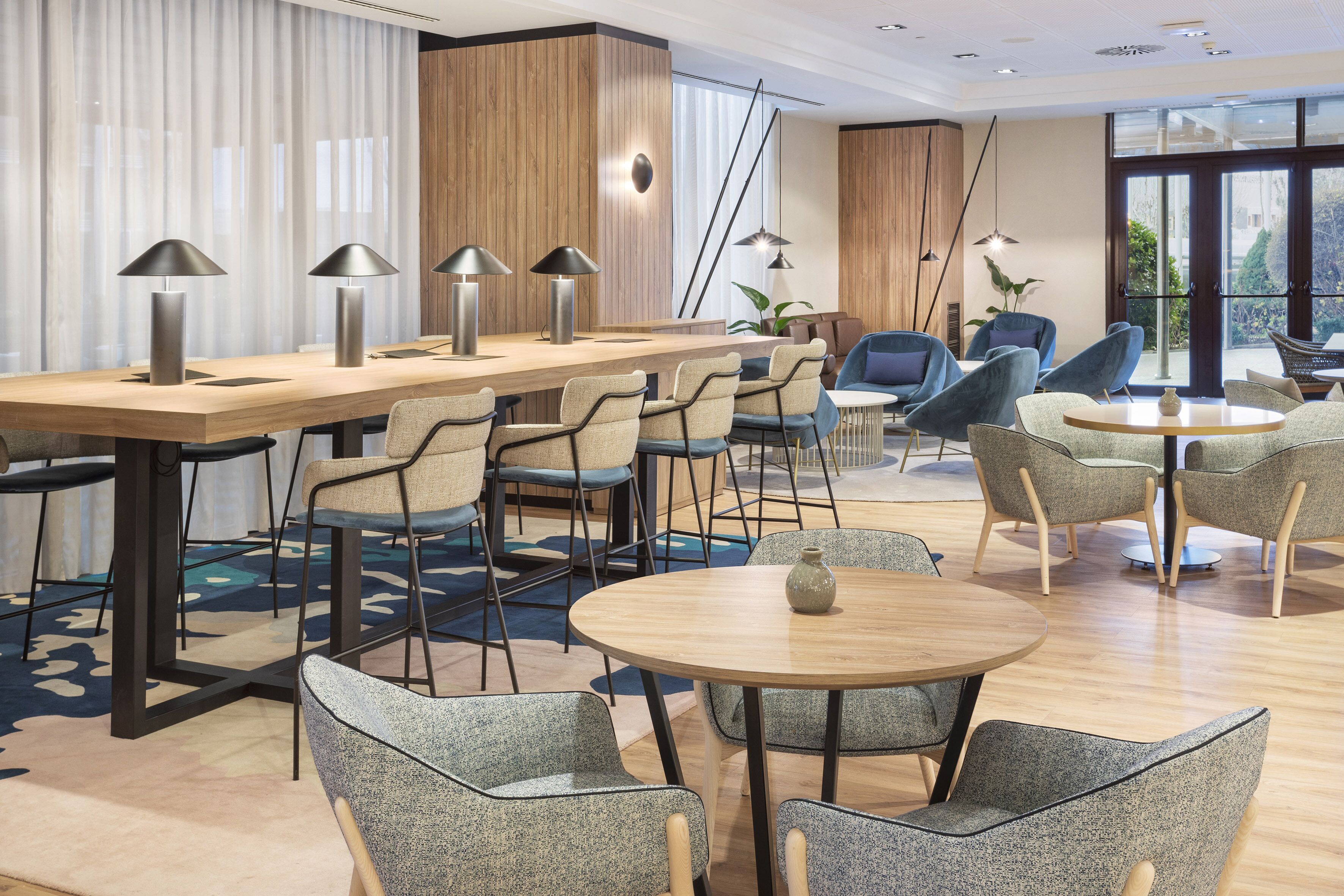Un moderno vestíbulo de hotel con un mostrador de recepción de madera, varias zonas de estar con sillas y mesas, y grandes ventanales que ofrecen vistas al exterior.