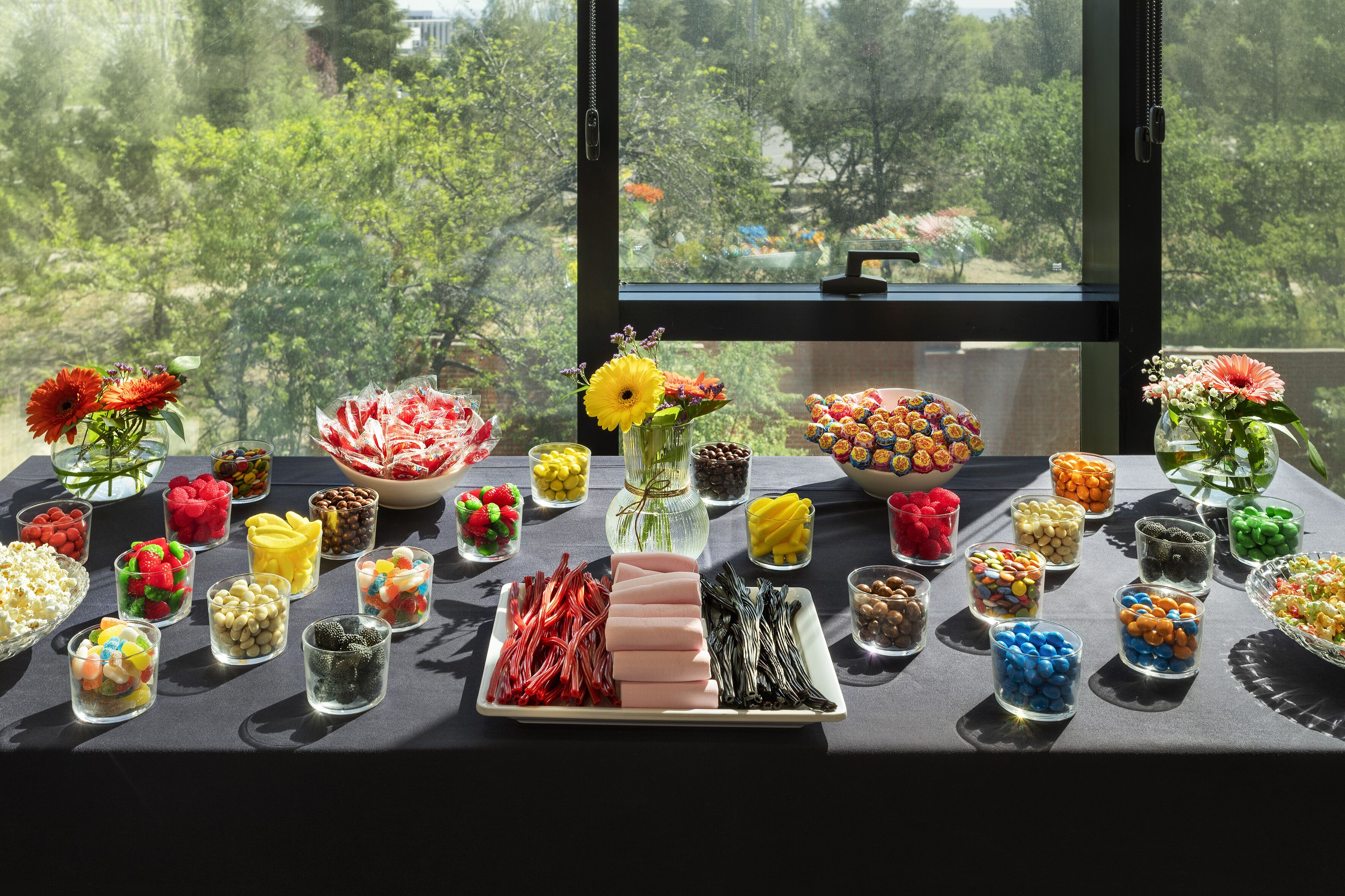Una mesa con diversos alimentos y flores, vista a través de una ventana que da a árboles y edificios.