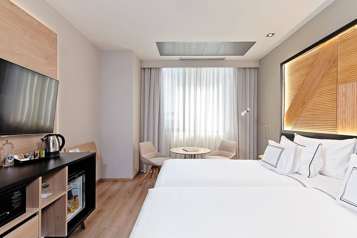 A hotel room with a bed, a desk with a kettle, a small table, two chairs, a TV, and a window with curtains.
