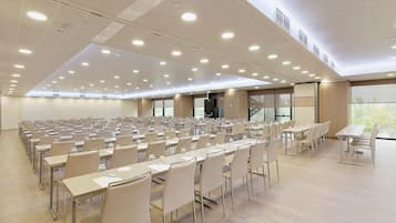 Une grande salle de conférence avec de nombreuses tables et chaises disposées en rangées, un plafond haut doté d’un éclairage encastré et de grandes fenêtres laissant entrer la lumière naturelle.
