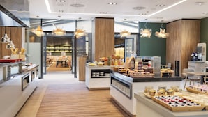 A modern cafe with wooden flooring, a variety of food displays, and pendant lighting. - Melia Avenida América (Madrid)