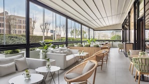 A modern outdoor seating area with white sofas and wicker chairs, large windows offering views of buildings and trees, and a tiled floor. - Melia Avenida América (Madrid)