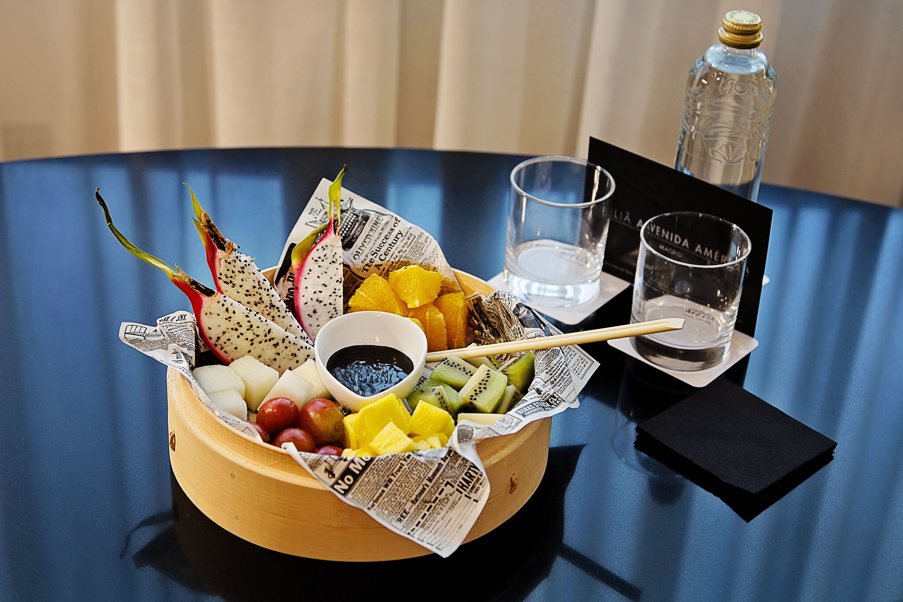 Un cuenco de madera lleno de frutas y verduras variadas, junto con un vaso de agua y un vaso pequeño, sobre una mesa con mantel azul.