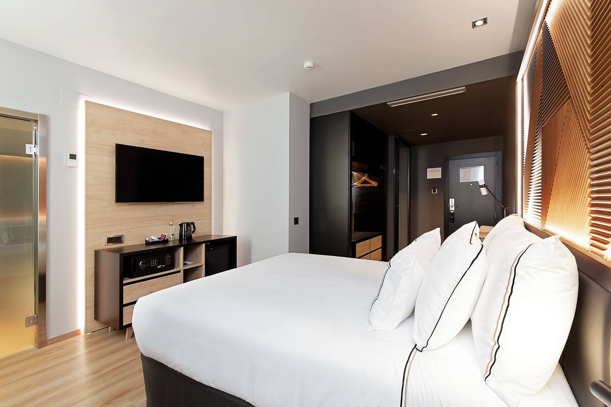 A modern hotel room with a large bed, a flat-screen TV mounted on the wall, a desk with a computer, and a walk-in closet.