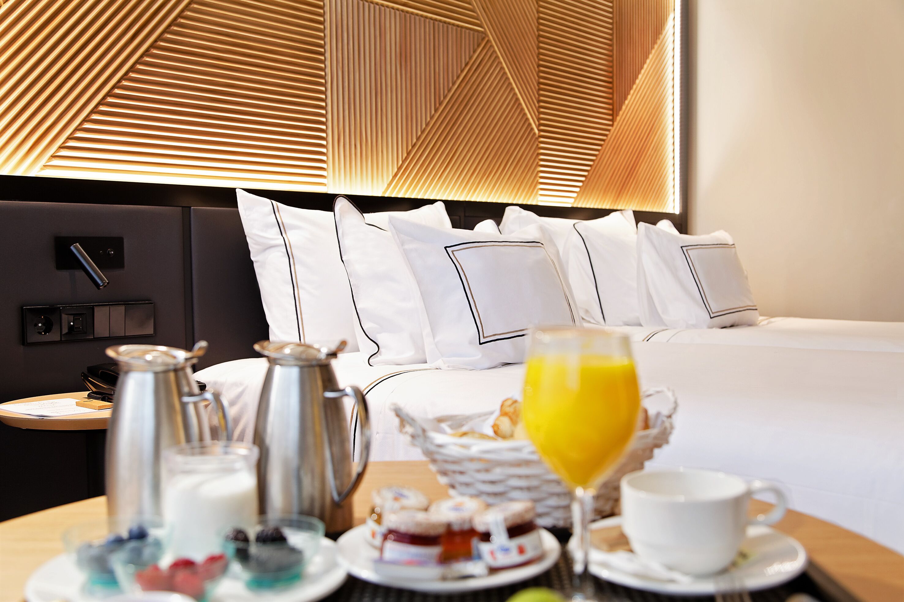 A neatly arranged breakfast table with a coffee pot, milk jug, and pastries, set against a modern room with a geometric patterned ceiling and a neatly made bed with white linens.