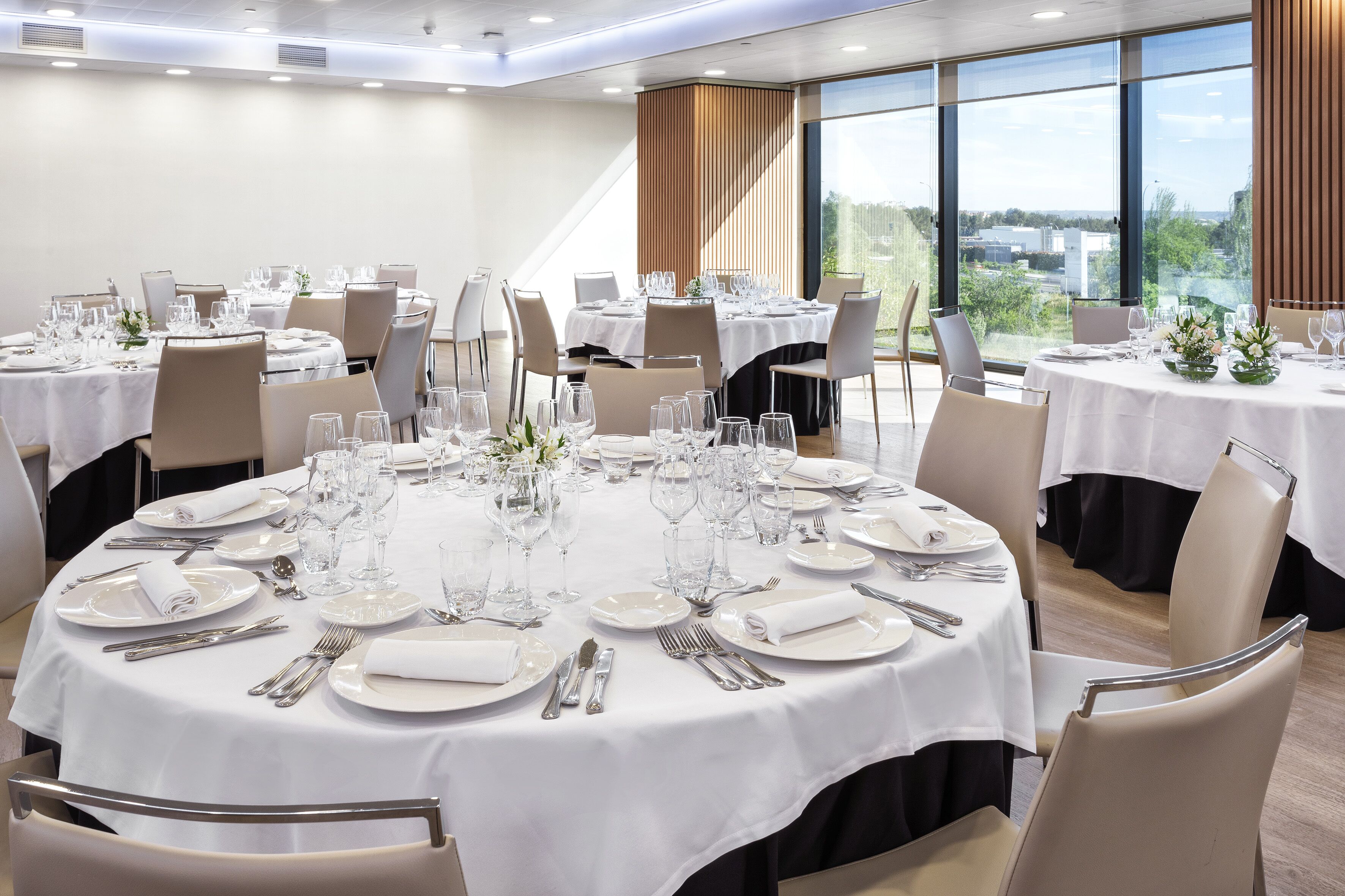 Un comedor con mesas redondas dispuestas para varios comensales, con manteles blancos, cubiertos ordenados y copas.