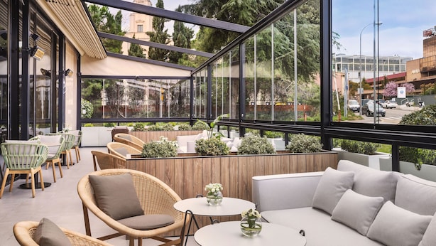 A modern outdoor seating area with wicker furniture, round tables, and a view of the cityscape through large windows. - Melia Avenida América (Madrid)