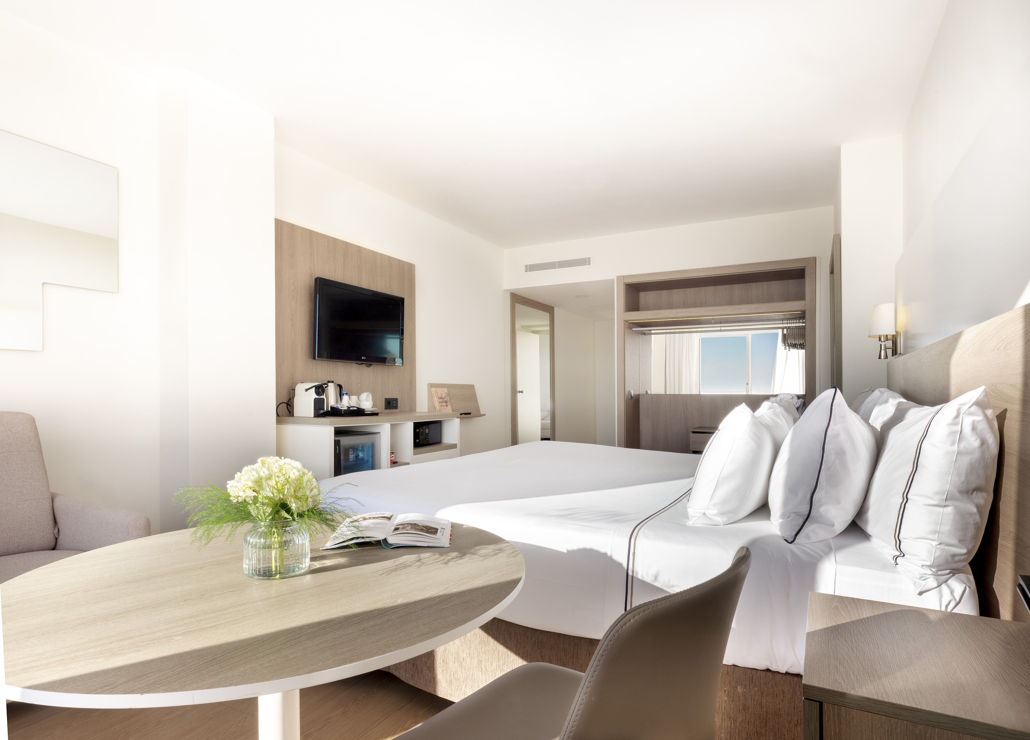 Una habitación de hotel moderna con una cama grande, un escritorio con silla, un televisor montado en la pared y un ventanal con cortinas.