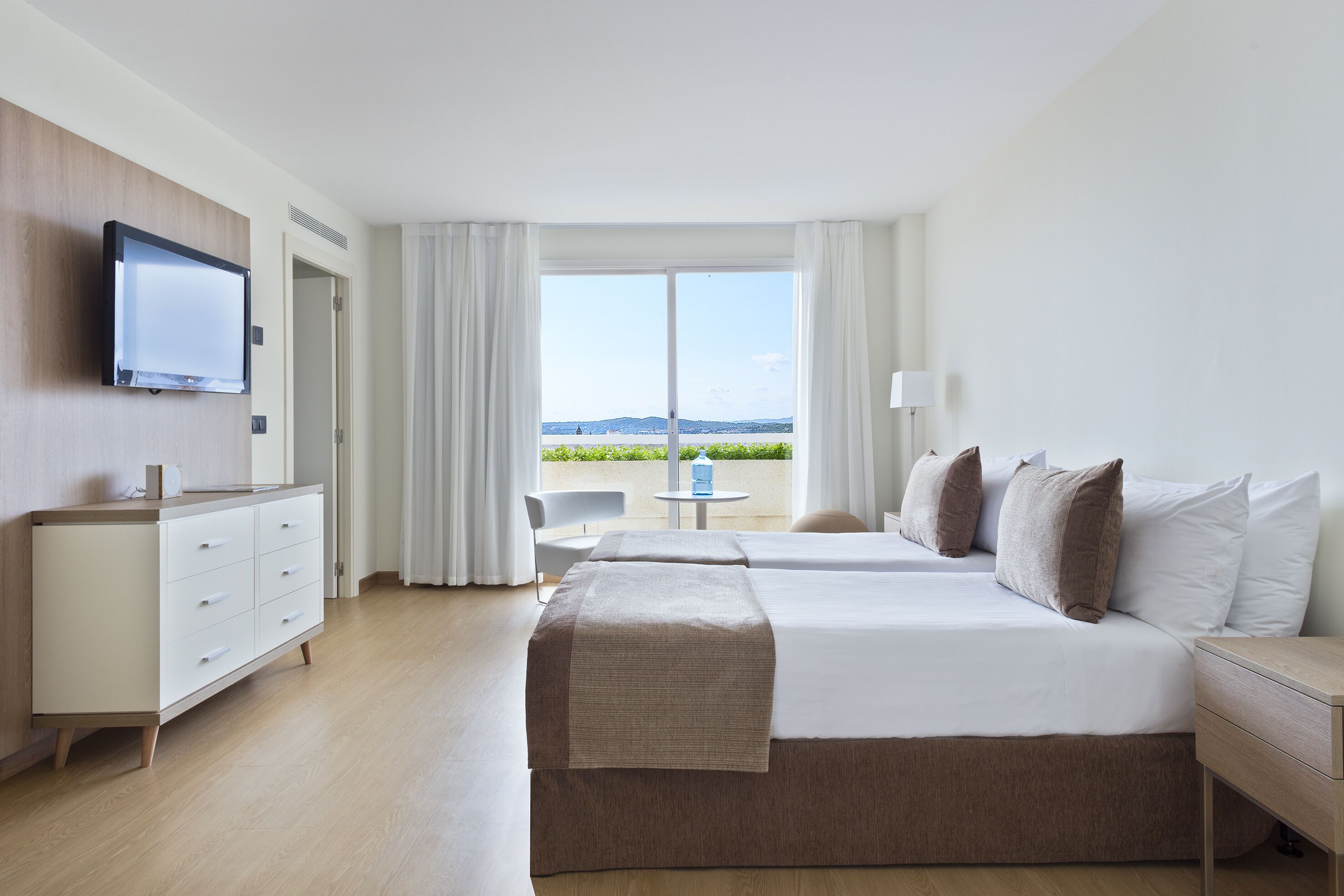 A hotel room with a bed, bedside table, TV, and a balcony with a view.