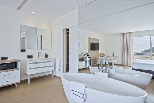 A modern hotel room with a large bathtub, a vanity with a mirror, a television, and a view of the outdoors.
